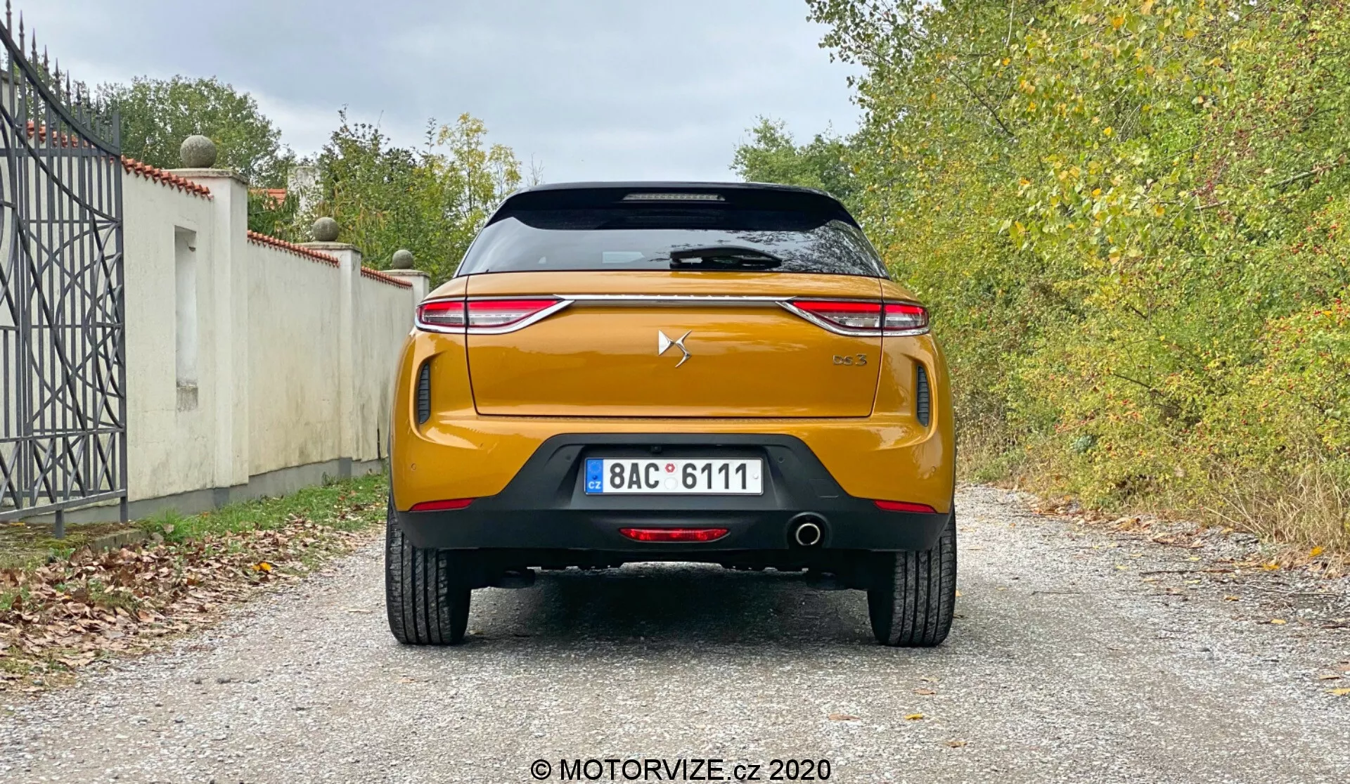 Pohľad zozadu na moderný automobil DS-Automobiles DS-3 Crossback 2019 s dizajnovými a technologickými prvkami, vrátane strešného spojlera, zadného okna s vykurovacím prvkom, vysoko montovanej brzdy, zadného krytu batožinového priestoru s logom výrobcu, výrazného chrómového pásu, elegantných LED zadných svetiel, robustného zadného nárazníka s parkovacími senzormi, dvojitého výfukového systému, čierneho difúzorového prvku a výraznej oranžovej alebo medenej farby na štrkovom povrchu s vegetáciou v pozadí.