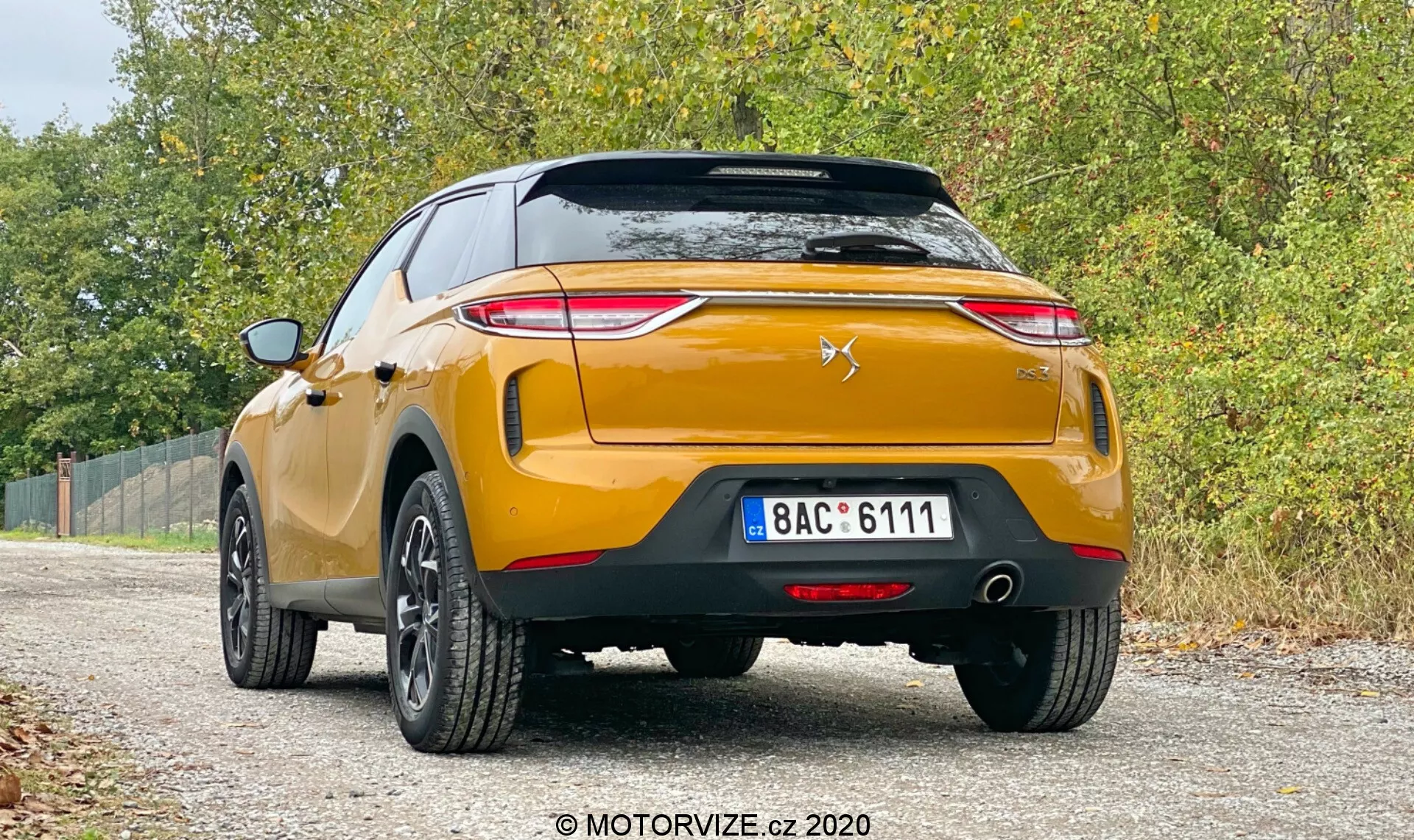 Obrázok ukazuje zadný pohľad na moderné, zlaté DS-3 Crossback 2019, zaparkované na neupravenej krajnici s vegetáciou po oboch stranách. Auto, zachytené z nízkeho uhla, zobrazuje ostré koncové svetlá smerujúce k stredu kufra, štýlový znak chevron a leva na zadných dverách, robustný zadný nárazník s parkovacími senzormi, dvojité výfuky, čierne plastové lišty a päťlúčové zliatinové kolesá. EČV je európskeho typu s bežnými zadnými odrazkami a zadným hmlovým svetlom. Mäkké, rozptýlené osvetlenie naznačuje zamračený deň, čo pridáva k neformálnemu, všestrannému prostrediu, zatiaľ čo vodoznak ‘MOTORVIZE.cz 2020’ naznačuje zdroj obrázku.
