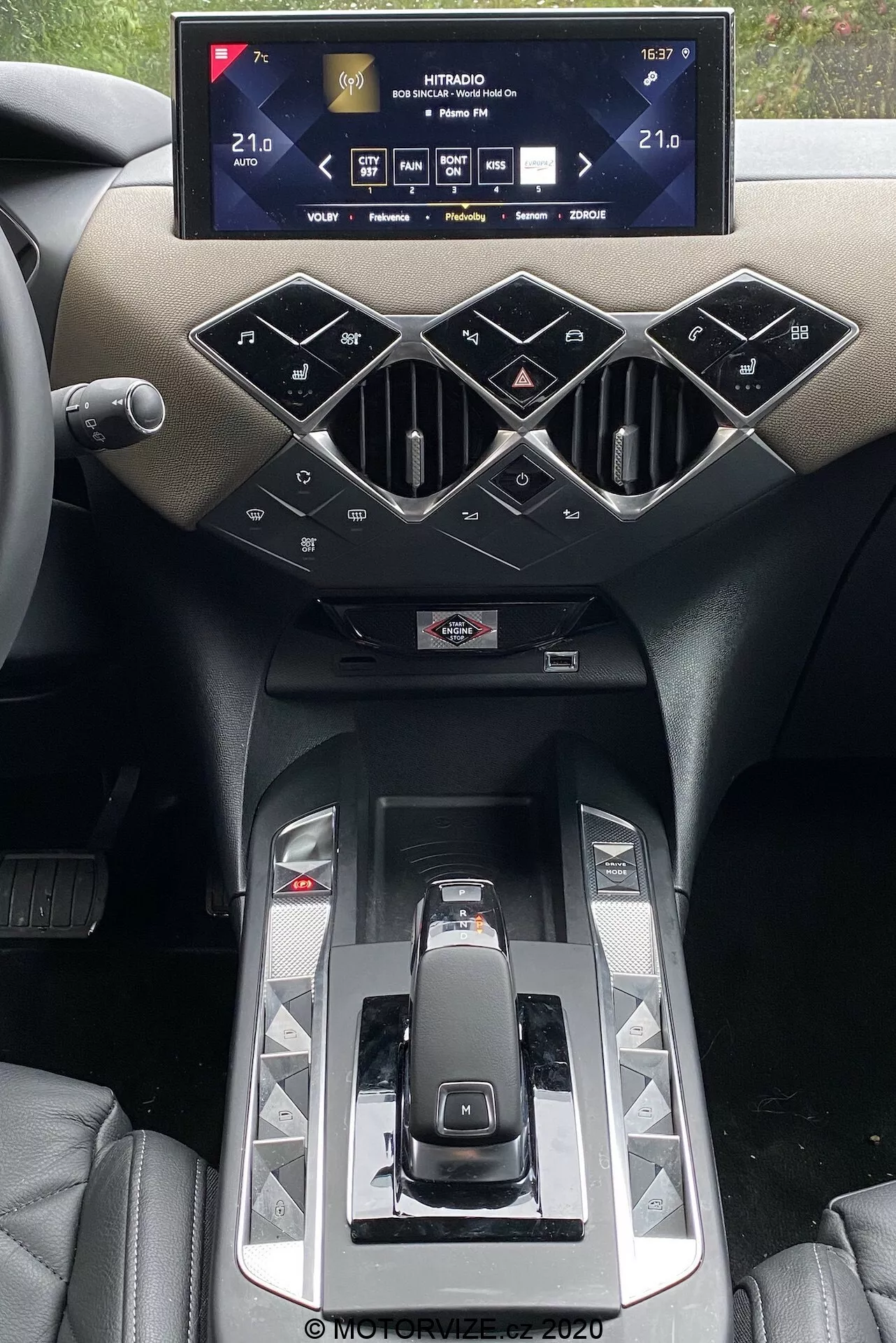 Interiér DS Automobiles DS 3 2019 zameraný na stredovú konzolu s infotainment systémom. Široký, horizontálne orientovaný dotykový displej zobrazuje nastavenia vozidla, vrátane teplôt klimatizácie nastavených na 21,0 °C. Pod ním sú geometricky tvarované vetracie otvory a fyzické tlačidlá v diamantovom usporiadaní, ktoré ovládajú systémy auta. Moderná radiaca páka, lesklé panely a náznak držiakov na poháre sú zdôraznené luxusnými materiálmi a prémiovým dizajnom.