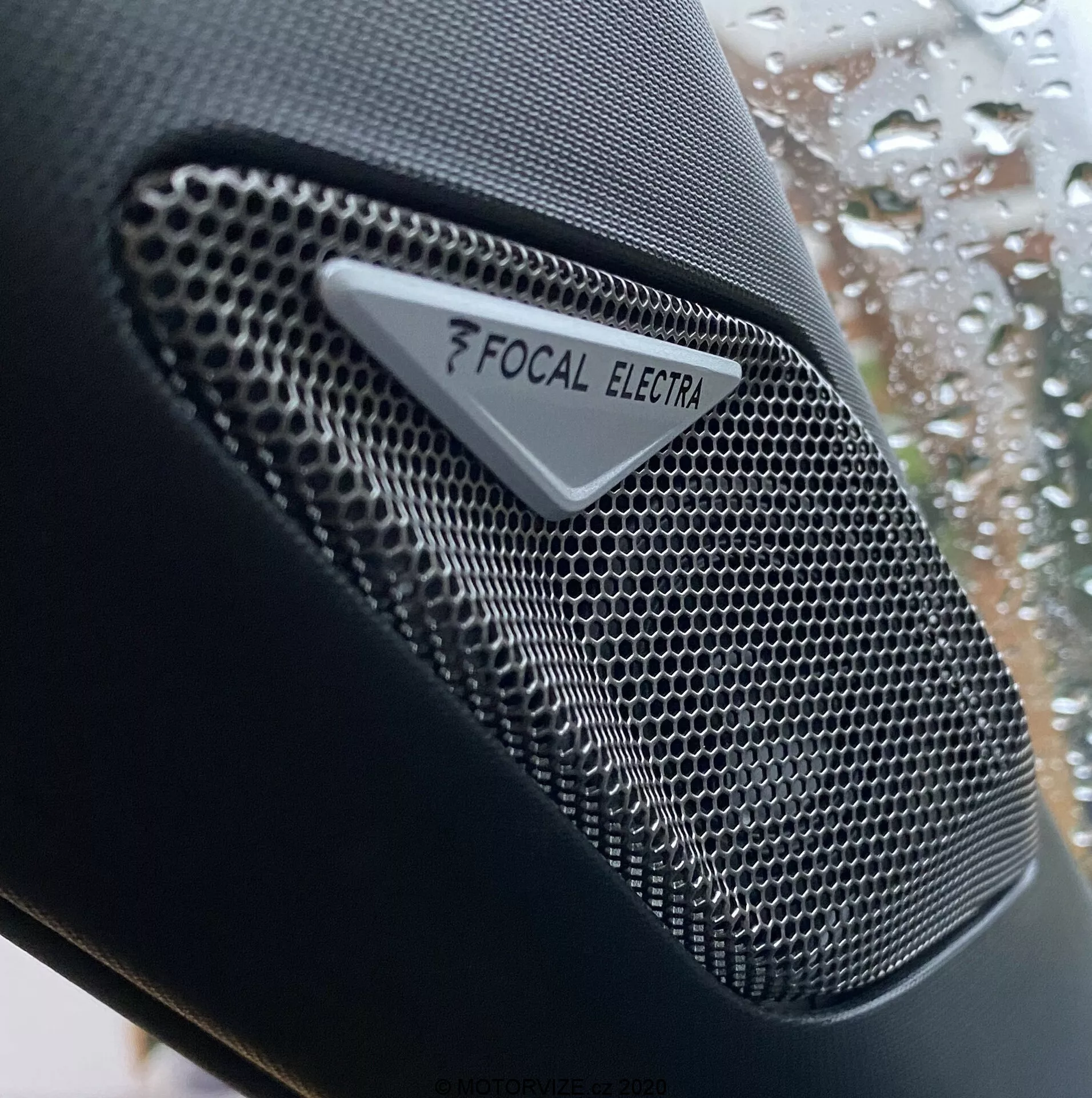 Detailný záber na reproduktor značky Focal Electra v DS Automobiles DS-3 Crossback 2019, zobrazujúci vysokofrekvenčný audio komponent s kovovou mriežkou v tvare včelieho plástu a jasným logom Focal a značkou Electra. Záber sa zameriava na zložitý dizajn reproduktora, s plytkou hĺbkou ostrosti, ktorá zdôrazňuje textúru reproduktora proti rozmazanému pozadiu okna auta s kvapkami dažďa.