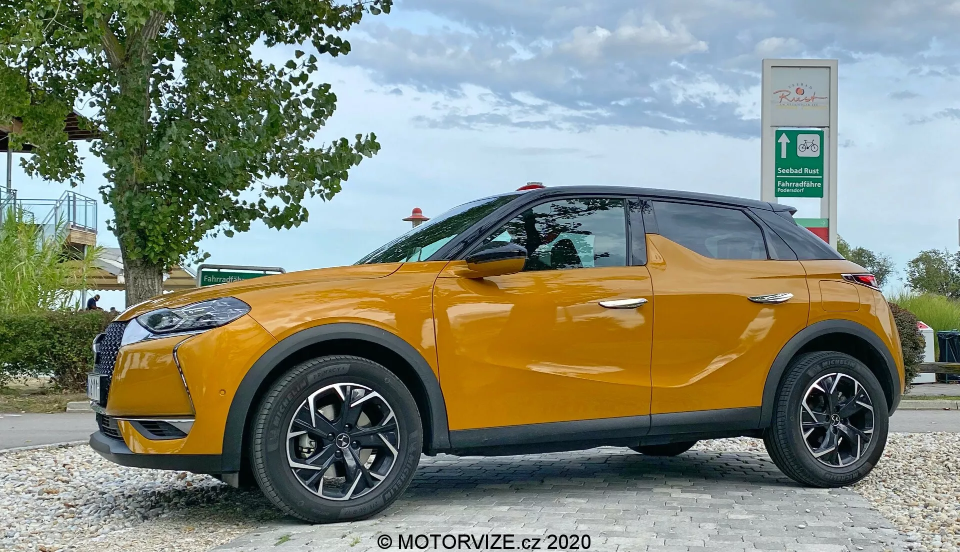 Bočný profil oranžového DS Automobiles DS 3 Crossback z roku 2019, zaparkovaného pri cestnej oblasti so zeleňou a značkou čerpacej stanice v pozadí. Strana vodiča je viditeľná s čiernymi viaclúčovými kolesami a zrkadlami v farbe karosérie z nízkeho uhla vo výške kolies auta, ukazujúc elegantný dizajn kompaktného SUV so stúpajúcou líniou okien a tónovanými sklami.