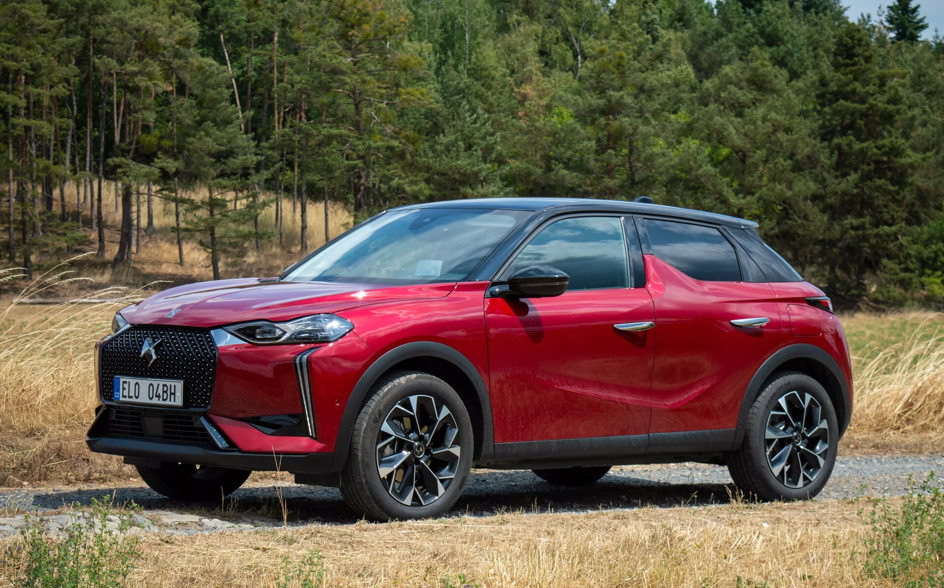 Bočný predný pohľad na DS Automobiles DS 3 E-TENSE 2023, červené SUV crossover, zliatinové kolesá s viacerými špicami, LED svetlomety, na štrkovej ceste s lesným pozadím
