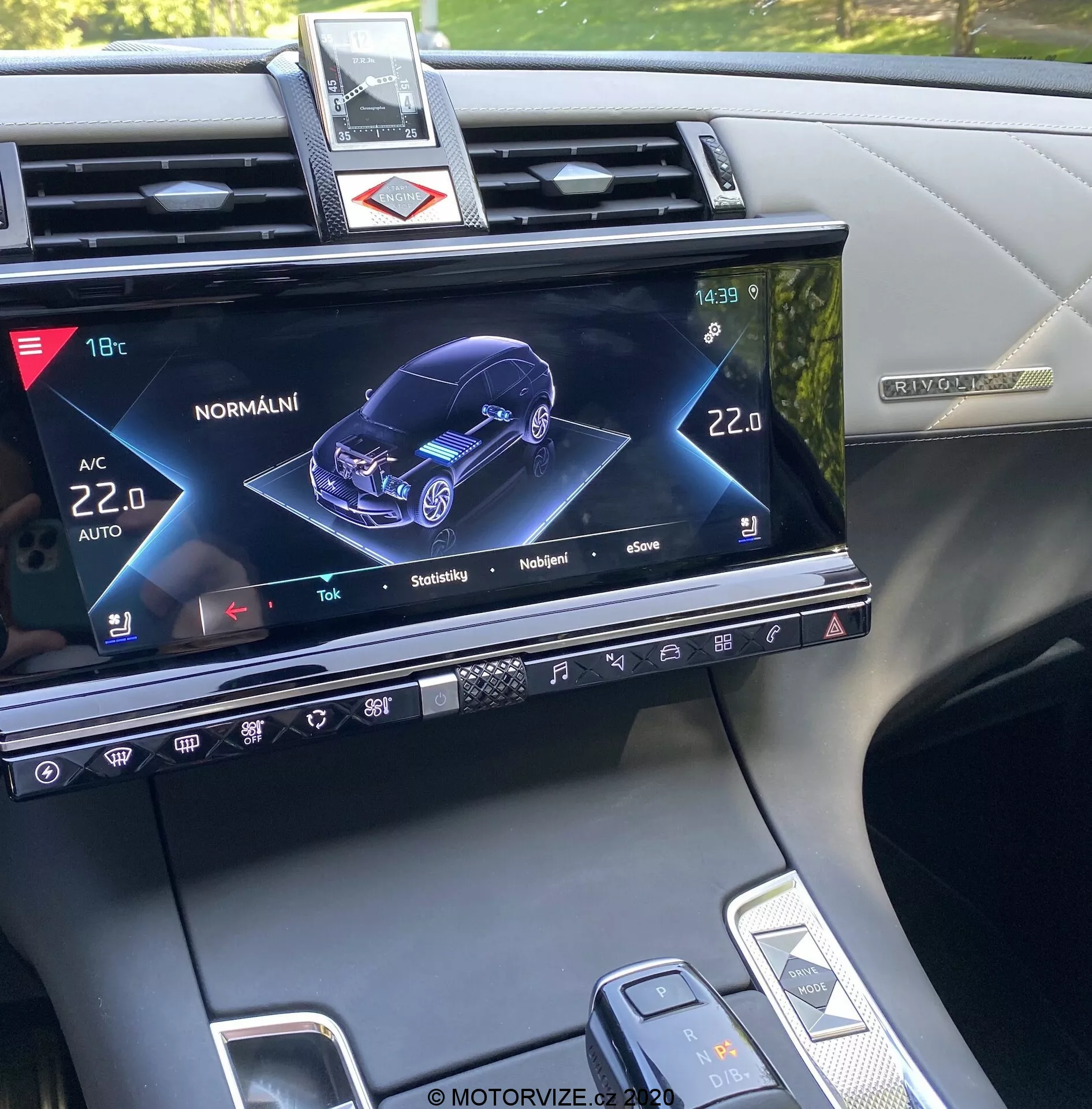 Centrálna konzola DS Automobiles DS-7 Crossback z roku 2018, zobrazujúca displej infotainment systému s 3D grafickým zobrazením auta z predného pravého uhla, ukazujúca jeho stav a rôzne nastavenia, ako je klimatizácia, vyhrievanie sedadiel a interaktívne funkcie modelu auta, spolu s fyzickými tlačidlami, dotykovými ovládacími prvkami a čiastočným pohľadom na radiacu páku a volič nastavení vozidla.