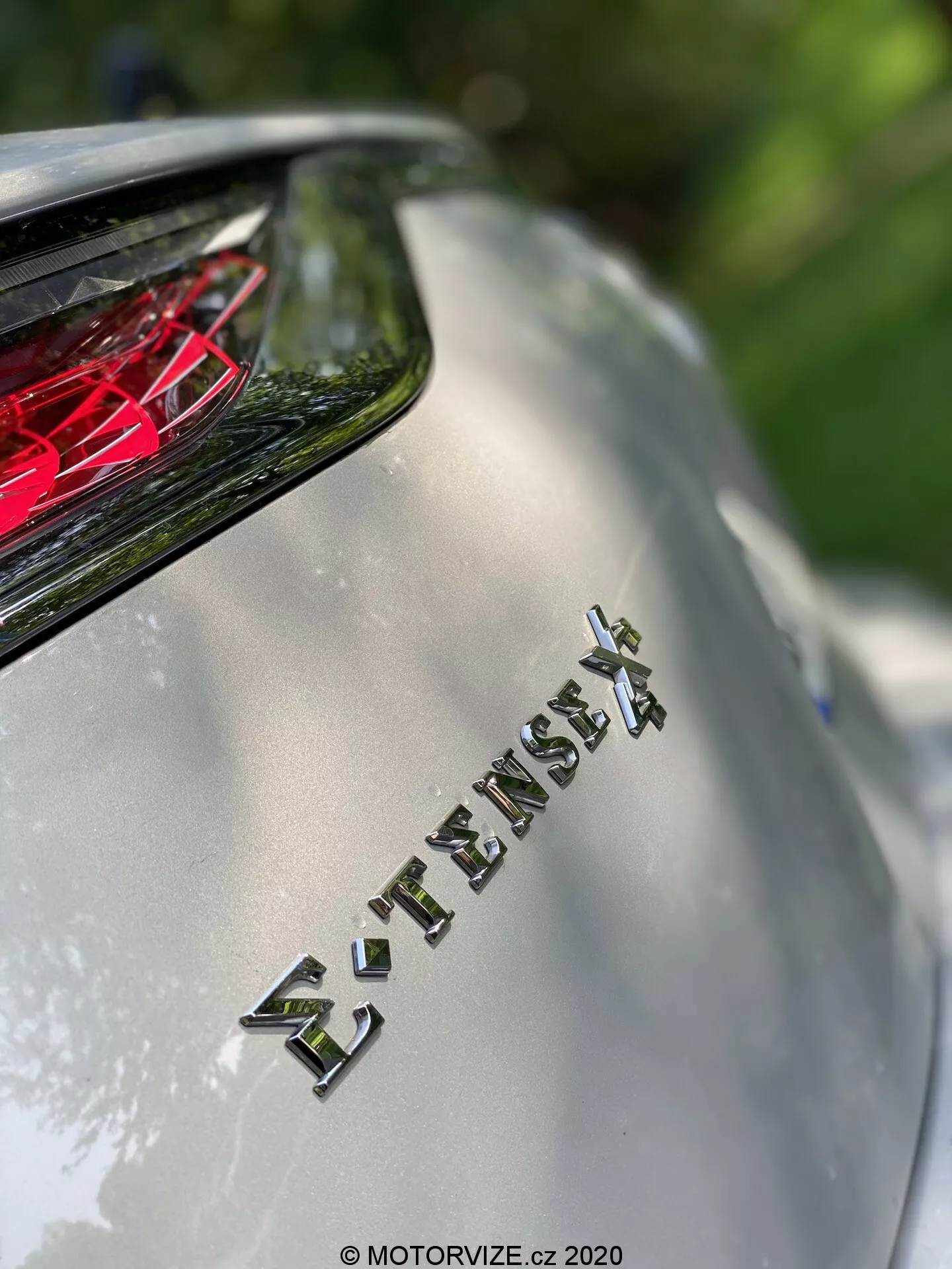 Detailný pohľad na zadnú časť modelu DS Automobiles DS 7 Crossback 2018, zameraný na znak 'E-TENSE 4X4', ktorý naznačuje pohon všetkých kolies a pravdepodobne hybridný alebo elektrický pohon. Plytká hĺbka ostrosti rozostruje pozadie a zvýrazňuje znak, nad ktorým je viditeľné červené kryštálové zadné svetlo. Obrázok zachytáva kovový povrch karosérie a chrómové označenie pod prirodzeným slnečným svetlom, zdôrazňujúci pokročilú technológiu a luxusný dizajn vozidla.