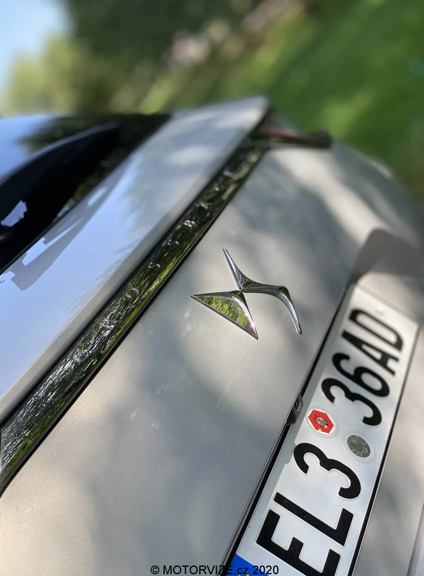 Detailný záber na zadnú časť strieborného DS Automobiles DS 7 Crossback z roku 2018. Obrázok zvýrazňuje kovový emblém auta umiestnený na strede zadného kufra, európsku poznávaciu značku s čiernymi znakmi 'EL3 36AD' pod emblémom a horný okraj zadného nárazníka. Viditeľná je tiež časť zadného skla s funkciou odhmlievania a vysoko lesklé reflexné povrchy. Plytká hĺbka ostrosti zdôrazňuje emblém, pričom jemne rozmazáva okraje kufra a zadného skla.