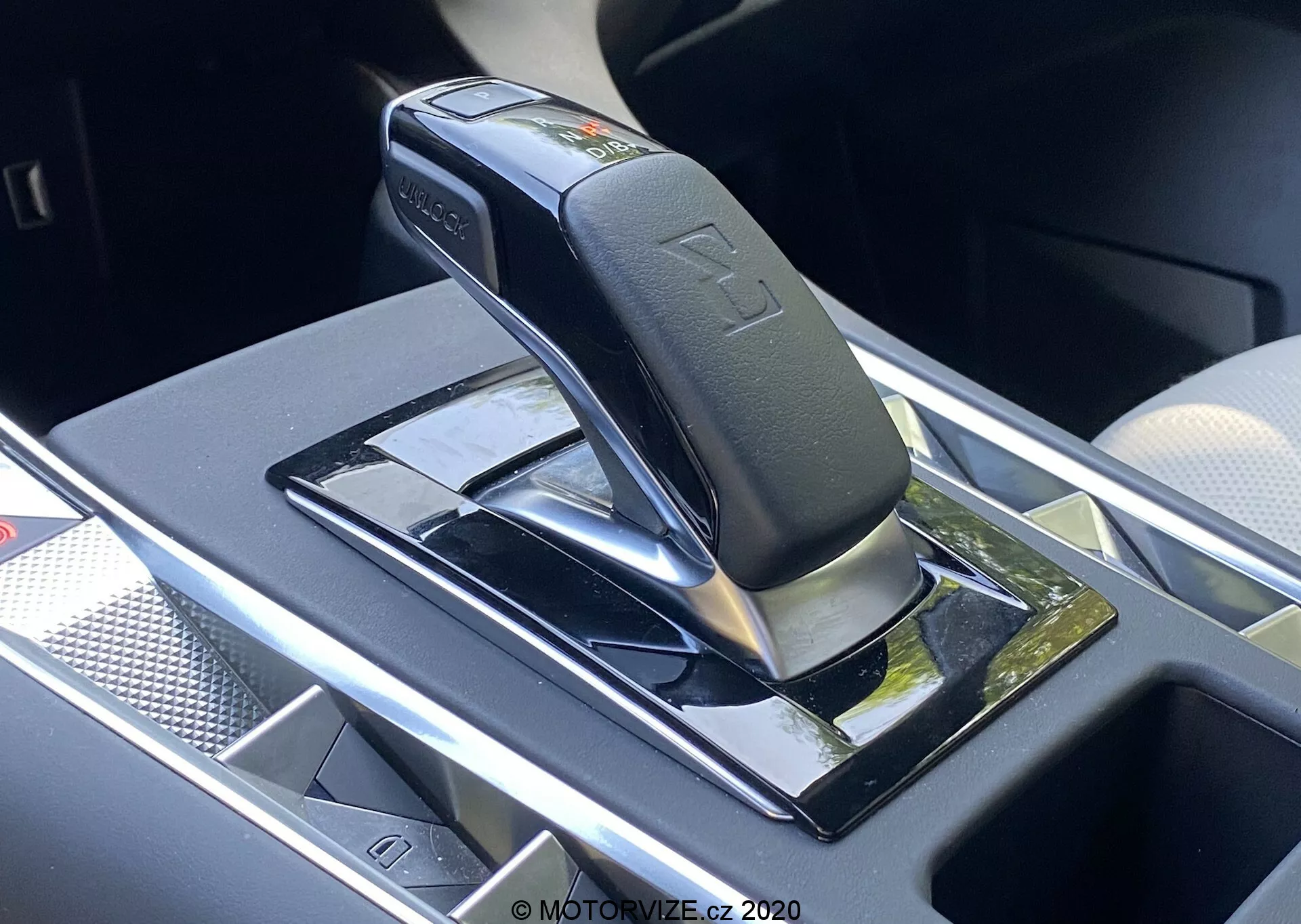 Zblízka pohľad na automatickú radiacu páku modelu DS Automobiles DS-7 2018 (DS-7 Crossback), zobrazujúci lesklú čiernu páku so striebornými akcentmi, pravdepodobne v parkovacej alebo neutrálnej polohe, namontovanú na klavírnom čiernom paneli s reflexnými povrchmi. Funkcie zahŕňajú vtlačené symboly prevodových polôh, bezpečnostné tlačidlo na páke a úložnú priehradku v blízkosti s potenciálnou bezdrôtovou nabíjačkou. Okolité oblasti majú chrómované alebo strieborné lemovanie, čo naznačuje prémiový, moderný interiér vozidla.