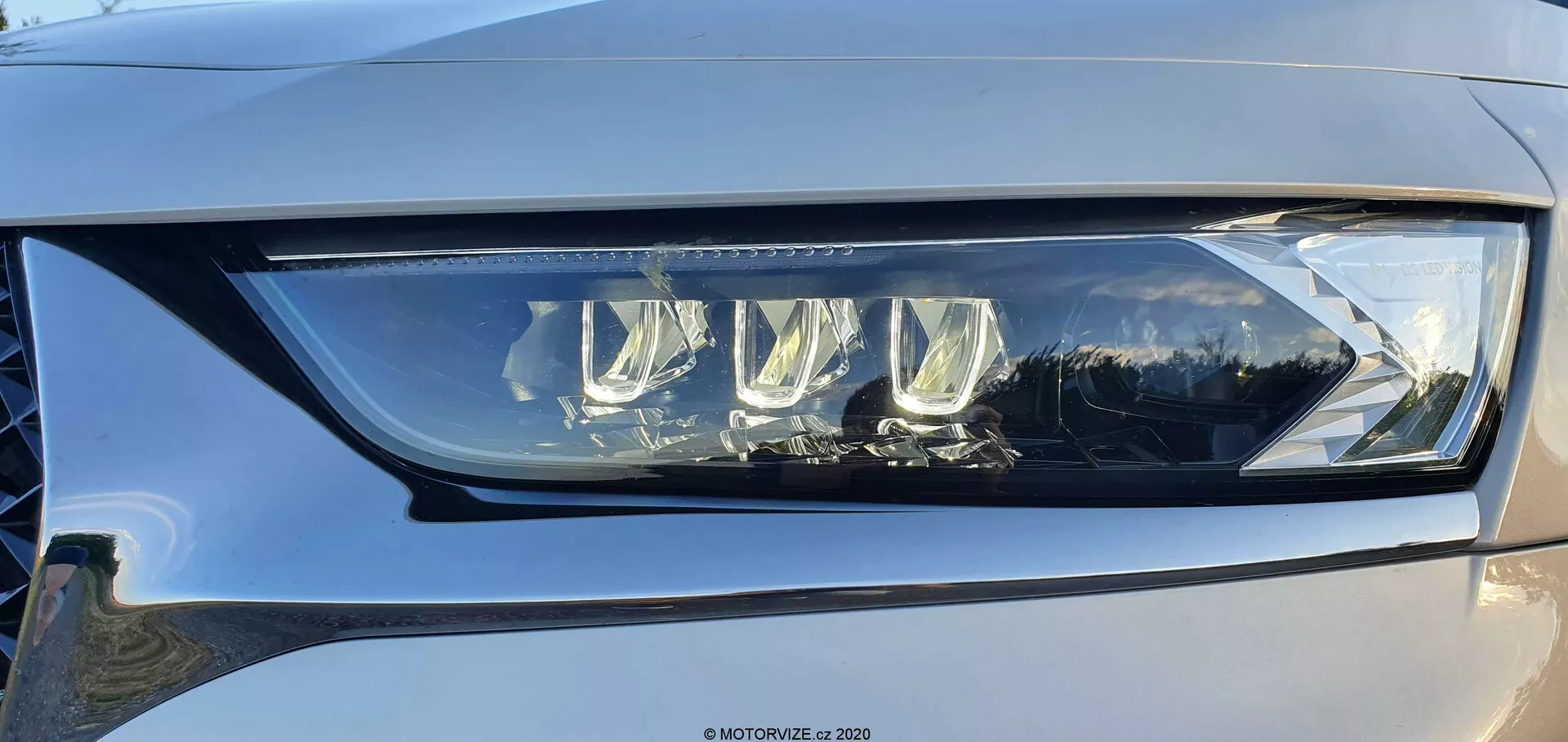 Detailný záber ľavej svetelnej jednotky DS Automobiles DS-7 Crossback z mierneho uhlovania, zobrazujúc moderný LED dizajn s viacerými projektorovými šošovkami a výrazným LED denným svietením zakapsulovaným v priesvitnom, vysokokvalitnom plaste. Svetlomet predstavuje pokročilú technológiu osvetlenia vozidiel schopnú poskytovať nízke aj vysoké svetelné lúče s možnými adaptívnymi funkciami pre lepšiu viditeľnosť. Viditeľný je jemný odraz na lesklom povrchu a náznak svetlo až stredne šedej farby vozidla.