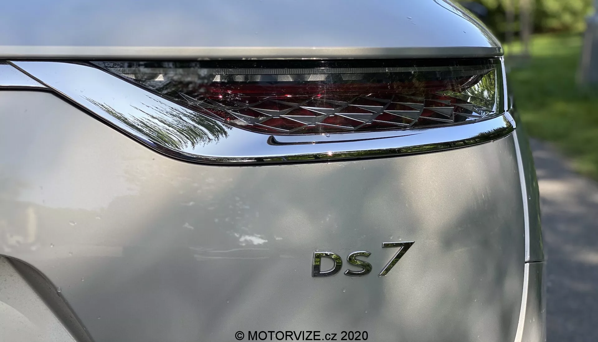 Zblízka zobrazenie zadnej časti modelu DS Automobiles DS 7 Crossback 2018 zhotovené z nízkeho uhla. Zameranie je na zadné dvere, na ktorých sa nachádzajú výrazné písmená 'DS' nad označením modelu '7'. Nad názvom modelu sa nachádza horizontálne rozšírené, zakrivené zadné svetlo s charakteristickými LED prvkami. Farba karosérie je metalická strieborná a sochárska dizajn naznačuje moderný a luxusný vzhľad. Fotografia zdôrazňuje branding a dizajn zadného svetla, s jemným odrazom jasného vonkajšieho prostredia na povrchu auta.