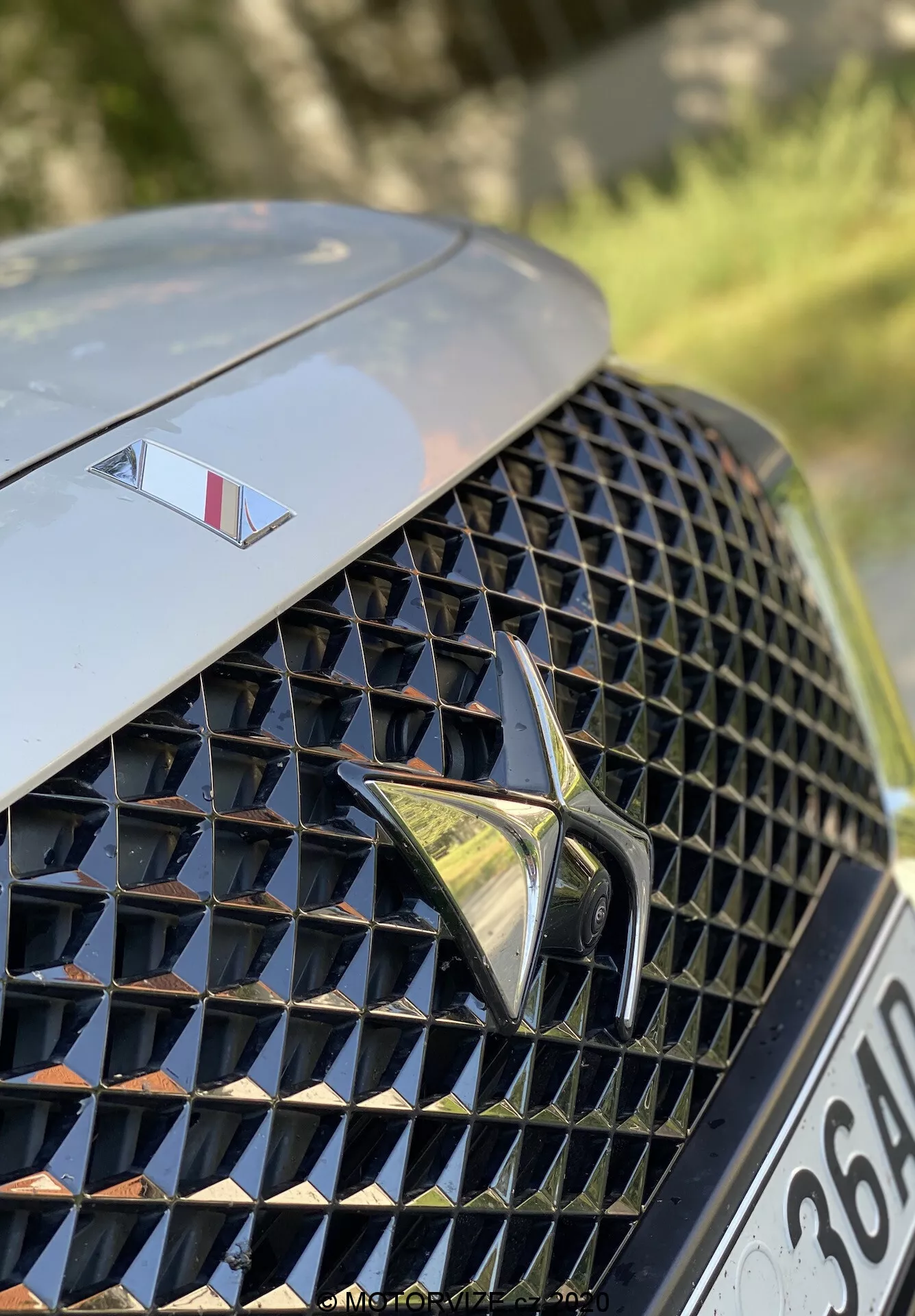 Detailný záber na prednú ľavú časť DS Automobiles DS 7 Crossback 2018. Obrázok sa zameriava na luxusnú, prepracovanú diamantovú mriežku s reflexnými povrchmi, uprostred so zvýrazneným štýlovým logom. Nad mriežkou je na kapote menší emblém, ktorý značí vyššiu triedu modelu. Pozadie je rozmazané, zvýrazňujúc dizajn mriežky a prvky značky.
