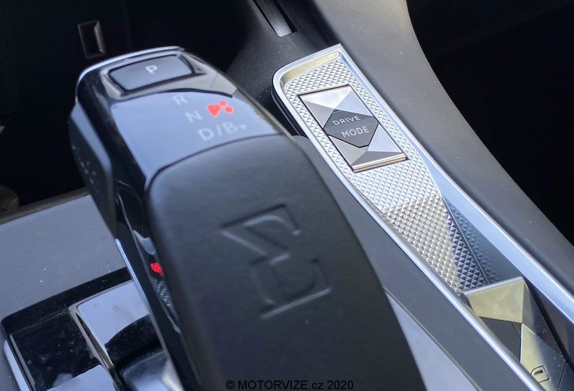 Detailný pohľad na stredovú konzolu modelu DS 7 Crossback 2018, zobrazujúci volič prevodovky a prepínač jazdných režimov z kosého uhla. Elektronický volič prevodovky v tvare L upozorňuje na polohu Park s červeným LED svetlom a tlačidlovou funkciou. Vpravo je prepínač jazdných režimov so vzorom diamantu a strieborným lemovaním, čo naznačuje prémiové prevedenie interiéru a pokročilé systémy riadenia vozidla.