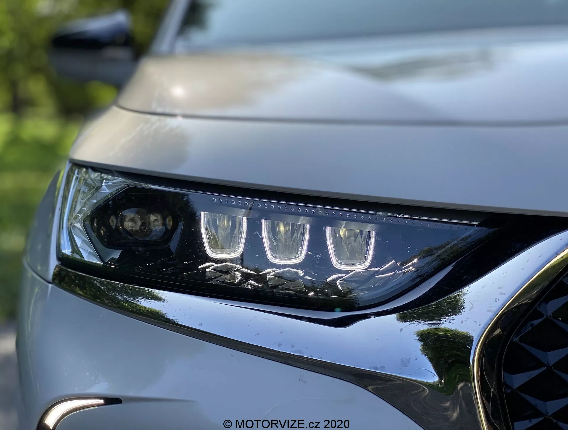 Detailný pohľad na ľavý svetlomet a prednú masku vozidla DS Automobiles DS 7 Crossback z roku 2018. Svetlomet má charakteristické LED denné svietenie a projektorovú šošovku pre hlavný lúč, obklopený viacerými LED prvkami. Elegantný a ostrý dizajn svetlometu ukazuje modernú aerodynamiku. Viditeľná časť tmavého, lesklého mriežky s kovovými akcentmi a časť tvarovanej kapoty zvýrazňujú dynamický, prémiový dizajn vozidla. Pozadie je rozmazané, čo zdôrazňuje ostrosť a detailné prevedenie svetlá a prednej časti vozidla.