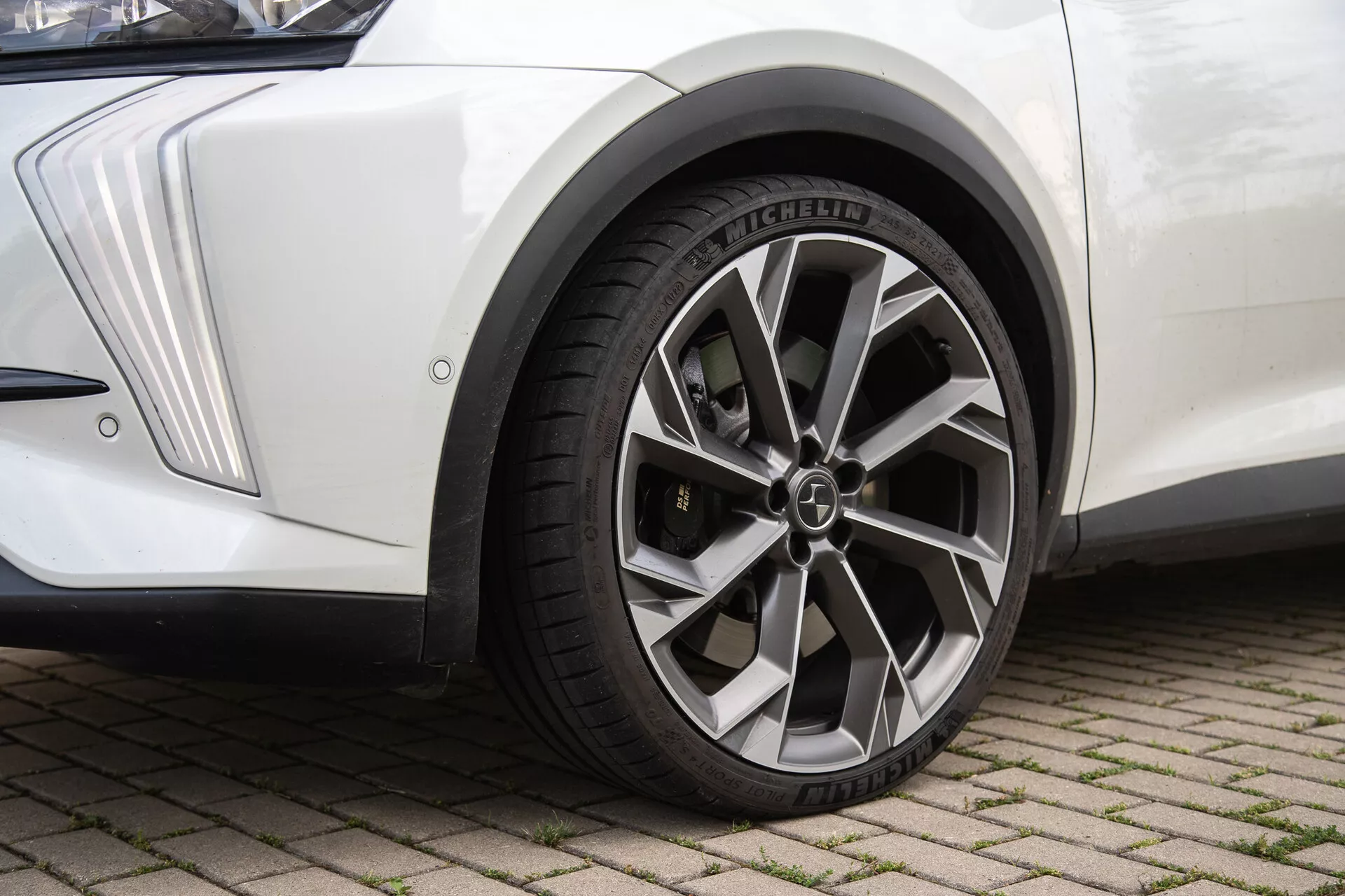 Detailný pohľad na predné koleso DS-Automobiles DS-7 2022 na dlažobnej ploche z prepojených tehál, s kovovým viackrúžkovým diskom kolies s krytom loga v strede, radiálnou pneumatikou Michelin s detailnými vzormi dezénu, bielym blatníkom s uhlovým dizajnom a čiernym striekacím štítom, pri prirodzenom osvetlení