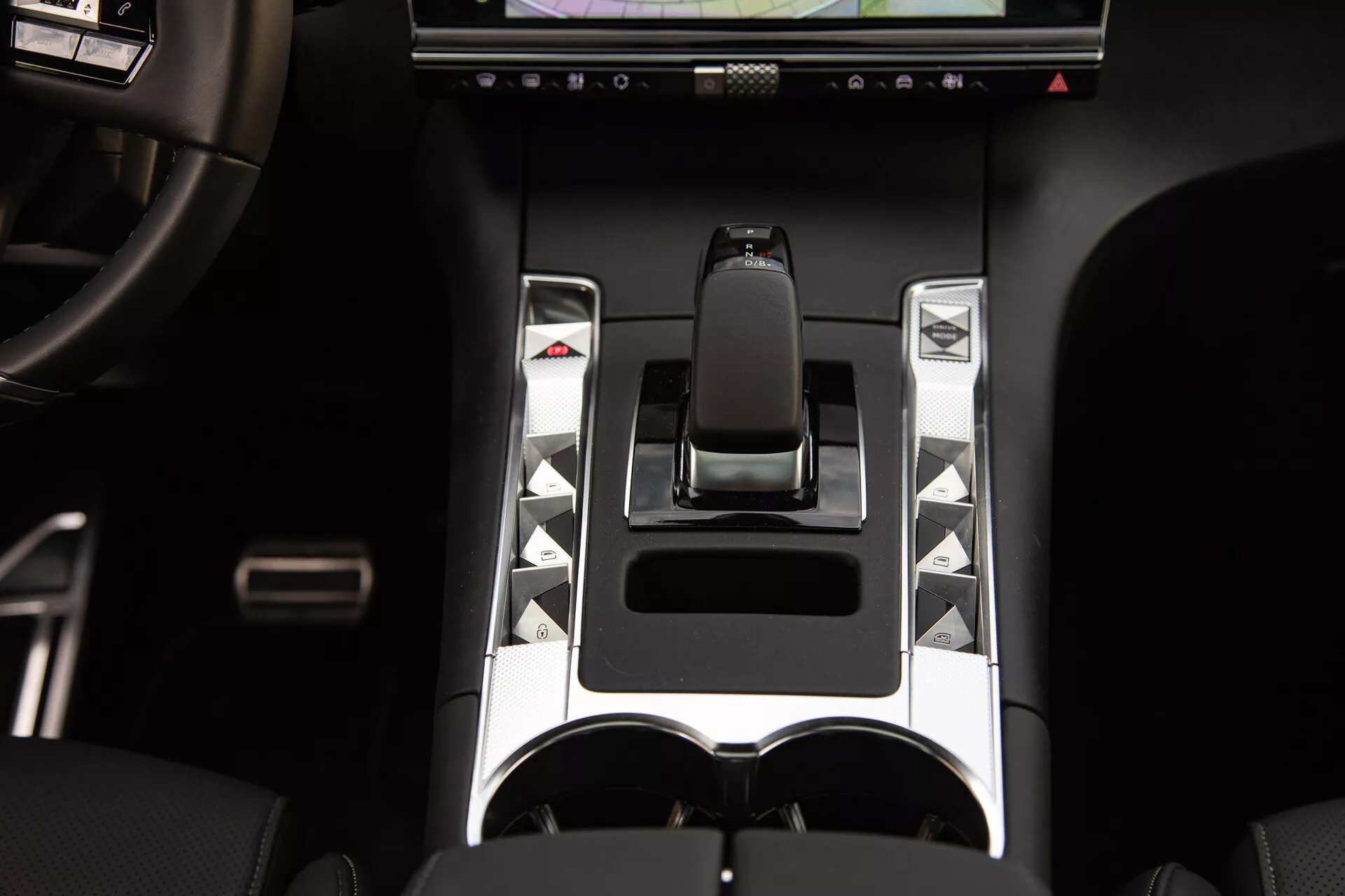Detailný pohľad na stredovú konzolu DS Automobiles DS-7 2022 s ergonomickou radicou pákou, kovovými akcentmi, unikátnymi ovládacími tlačidlami, dotykovým displejom a dvojitými držiakmi na poháre, ktoré zdôrazňujú pokročilé prvky ovládania vozidla a prvky luxusného dizajnu.