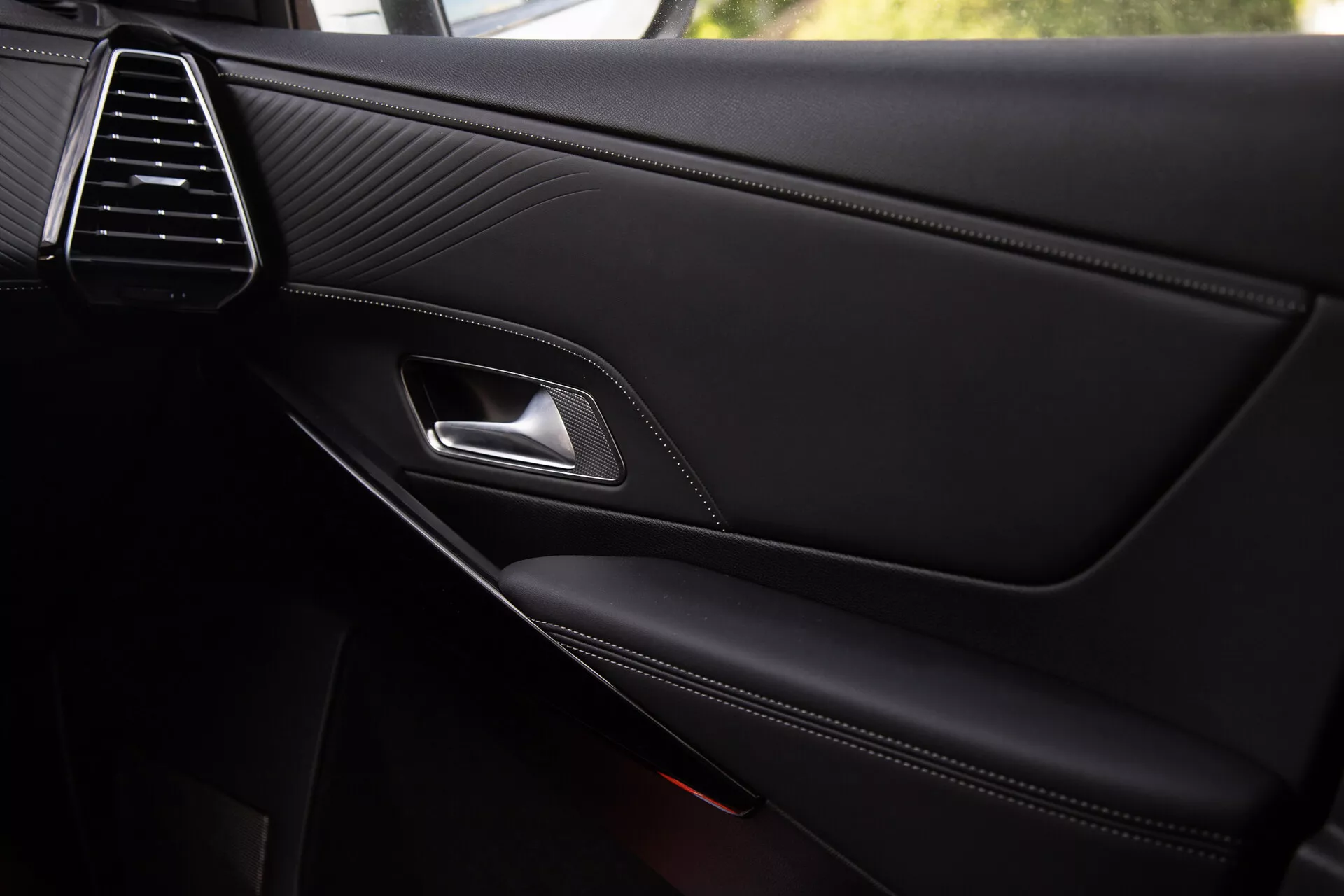 Detailný pohľad na vnútorný panel dverí DS Automobiles DS-7 2022 s elegantnou ventilačnou mriežkou, prešívanou kožou, chromovanou kľučkou dverí, ergonomickou páčkou zámku, kontrastne prešívanou opierkou a integrovaným úložným priestorom s čiastočne viditeľnou mriežkou reproduktora, odrážajúci prémiovú kvalitu a detaily dizajnu.