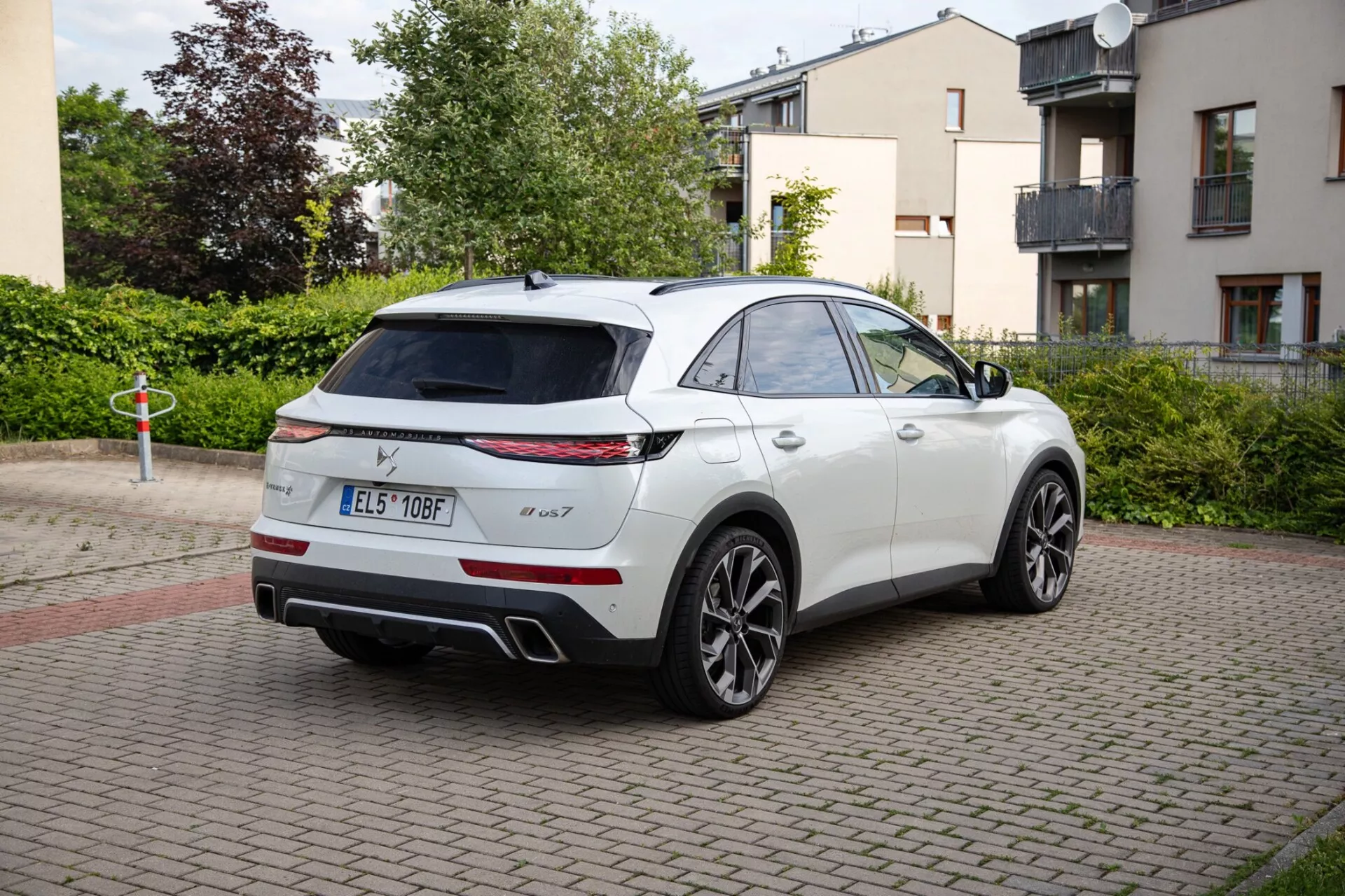 Biela SUV DS Automobiles DS-7 z roku 2022 zaparkovaná na oblasti s tehlovou dlažbou, zobrazujúca LED zadné svetlá, dvojitý výfukový systém a športové zliatinové kolesá. Obrázok zachytáva vozidlo so štýlovým dizajnom SUV pripomínajúcim kupé z trojštvrtinovej zadnej perspektívy, zdôrazňujúci možné panoramatické strešné okno a zrkadlá s technologickými vylepšeniami.