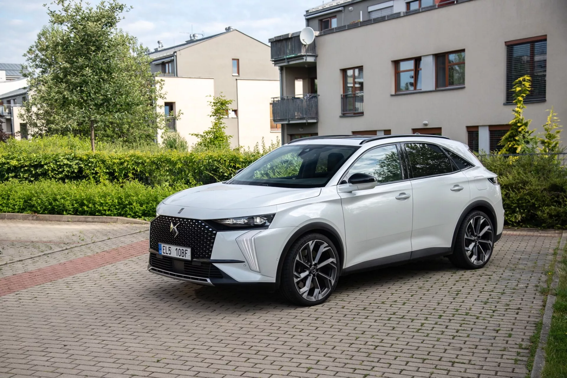 Trojštvrťový predný pohľad na crossover DS Automobiles DS-7 2022 zaparkovaný na obytnom ulici s dláždením z tehál, ktorý prezentuje jeho moderný dizajn vrátane výraznej prednej mriežky, štíhlych LED svetlometov, sochársky tvarovanej kapoty, veľkých zliatinových kolies a športového bočného profilu s integrovanými smerovými signálmi na dverách namontovaných bočných zrkadiel za denného prirodzeného svetla.