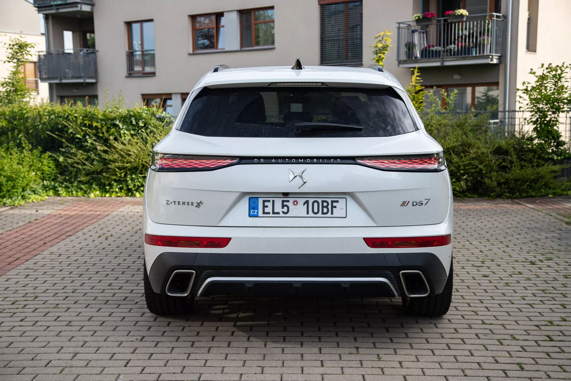 Zadný pohľad na zaparkovaný model DS Automobiles DS 7 2022 na tehlovej ploche s budovami a zeleňou na pozadí, predvádzajúci LED svetelný pás, zadné svetlá, dvojité výfukové otvory a EU evidenčnú značku