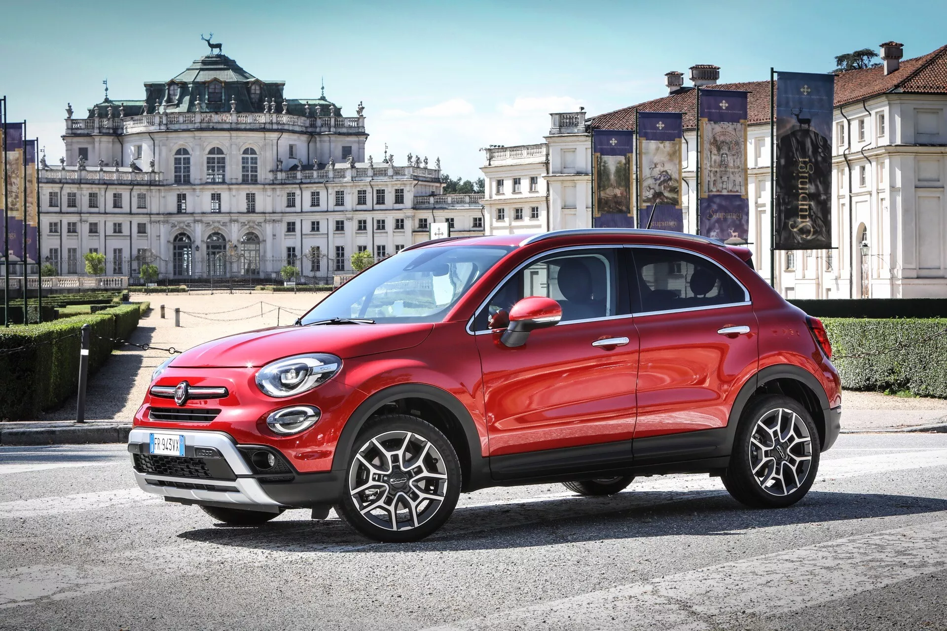 Trojštvrťový predný pohľad na červené SUV Fiat 500X (334-facelift) 2018, featuring a prominent grille with logo, round headlamps with daytime running lights, fog lamps on the front bumper with skid plate design indicating rugged capability, dynamic contour lines on the hood, high ground clearance, distinctive multi-spoke alloy wheels with black cladding on wheel arches for a sporty look, integrated turn signals on side mirrors, roof rails, chrome door handles and window outlines, zaparkované pod uhlom v mestskom prostredí s historickou architektúrou na pozadí