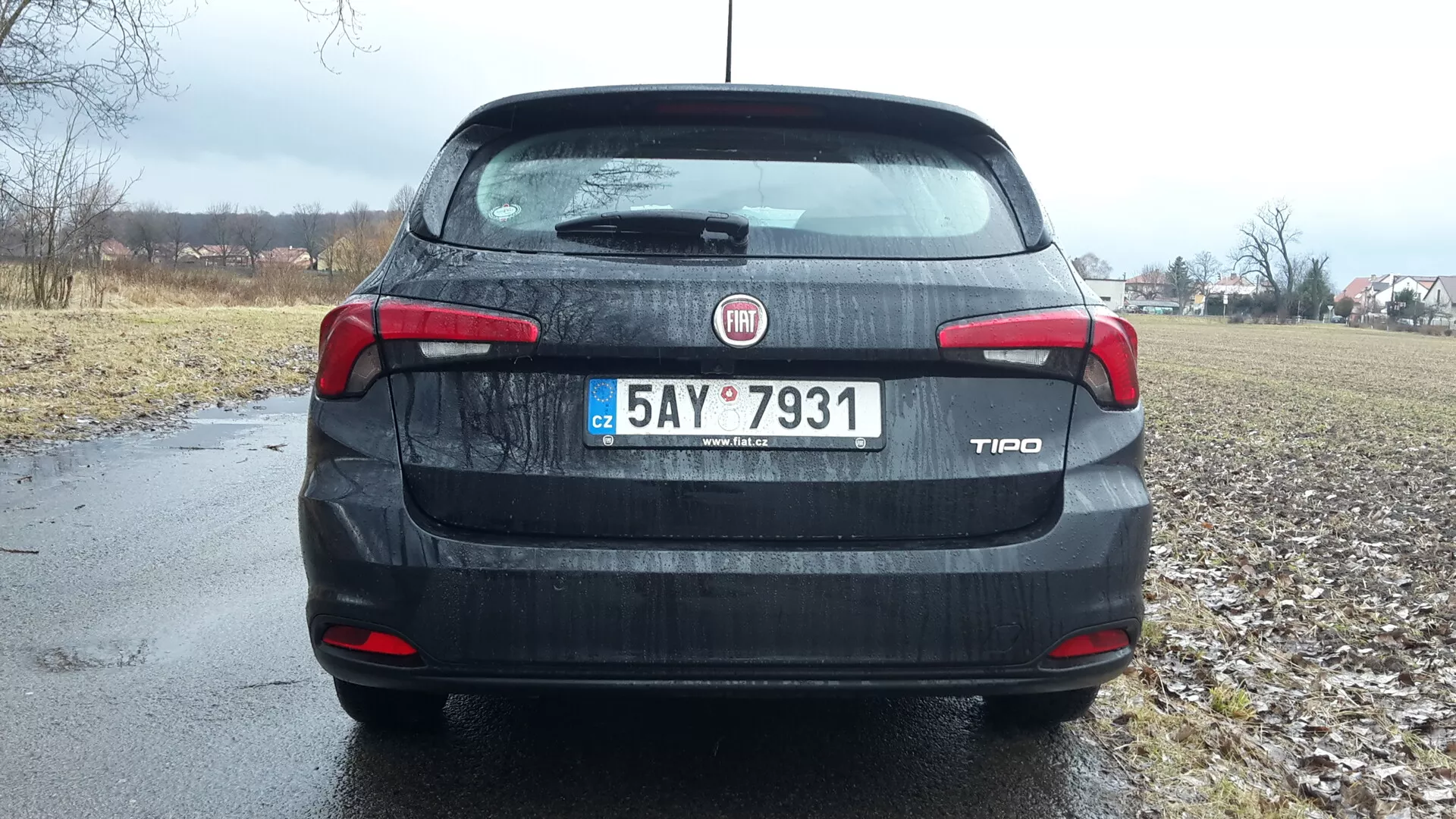 Zadný pohľad na sedan Fiat Tipo 2016 pod zamračenou oblohou, s identifikáciou značky a modelu na kufri, detailnými zadnými svetlami a tretím brzdovým svetlom, evidenčnou tabuľkou a náznakom vidieckeho prostredia na pozadí