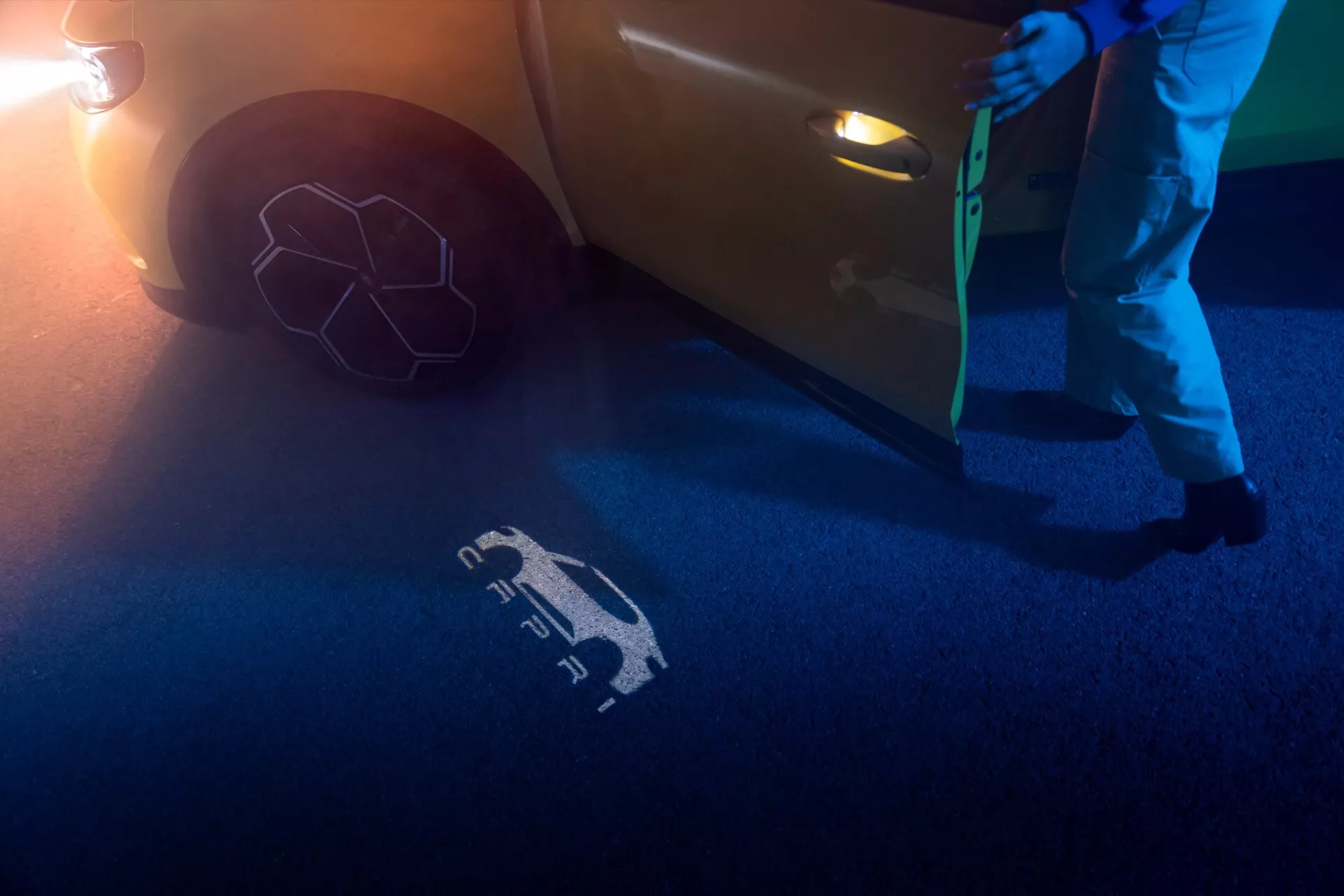 Čiastočný pohľad na Ford Capri 2024 z nízkeho uhla, zameraný na bok pri predných dverách, zobrazujúci spodnú časť predných dverí s modernou, zapustenou kľučkou a otvorenými dverami. Viditeľný je svetelný symbol premietaný na zem technológiou welcome/puddle light, osvetľujúci modré a oranžové odtiene v prostredí s nízkym osvetlením a nočnou atmosférou. Čiastočne je viditeľné predné koleso a pneumatika, s ďalším oslnením z jasného svetelného zdroja v pravom dolnom rohu.