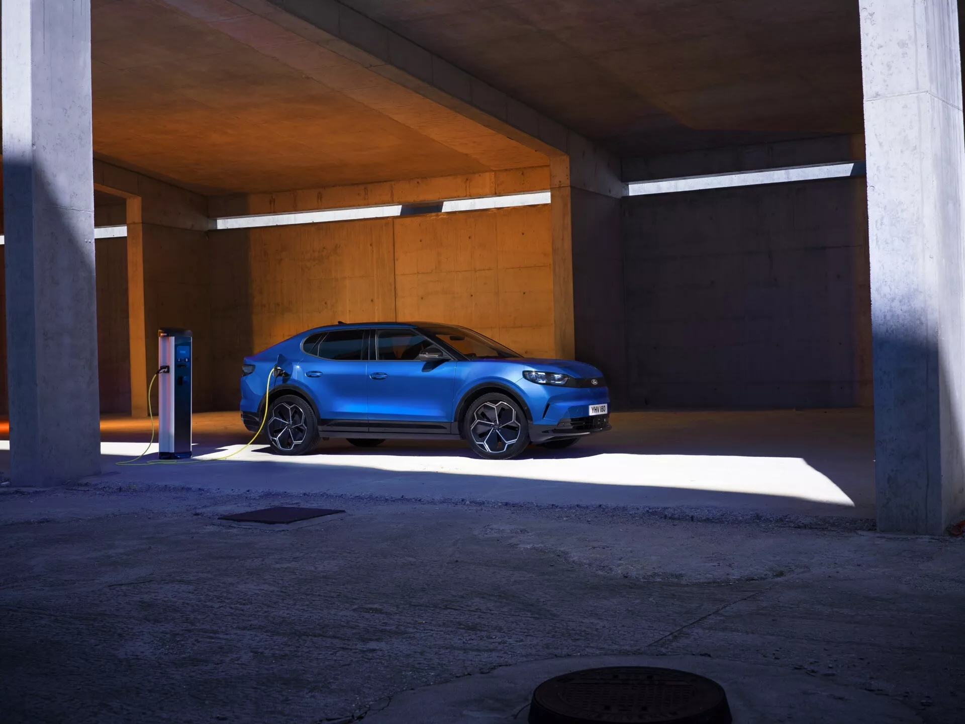 Žiarivo modré SUV Ford Capri 2024 zaparkované v betónovej parkovacej štruktúre, zobrazené z trojštvrťového predného pohľadu, ukazujúce jeho uhlové LED svetlomety, charakteristický dizajn masky a pripojenú nabíjaciu stanicu elektromobilov, zdôrazňujúc jeho moderný, športový dizajn a elektrické schopnosti.