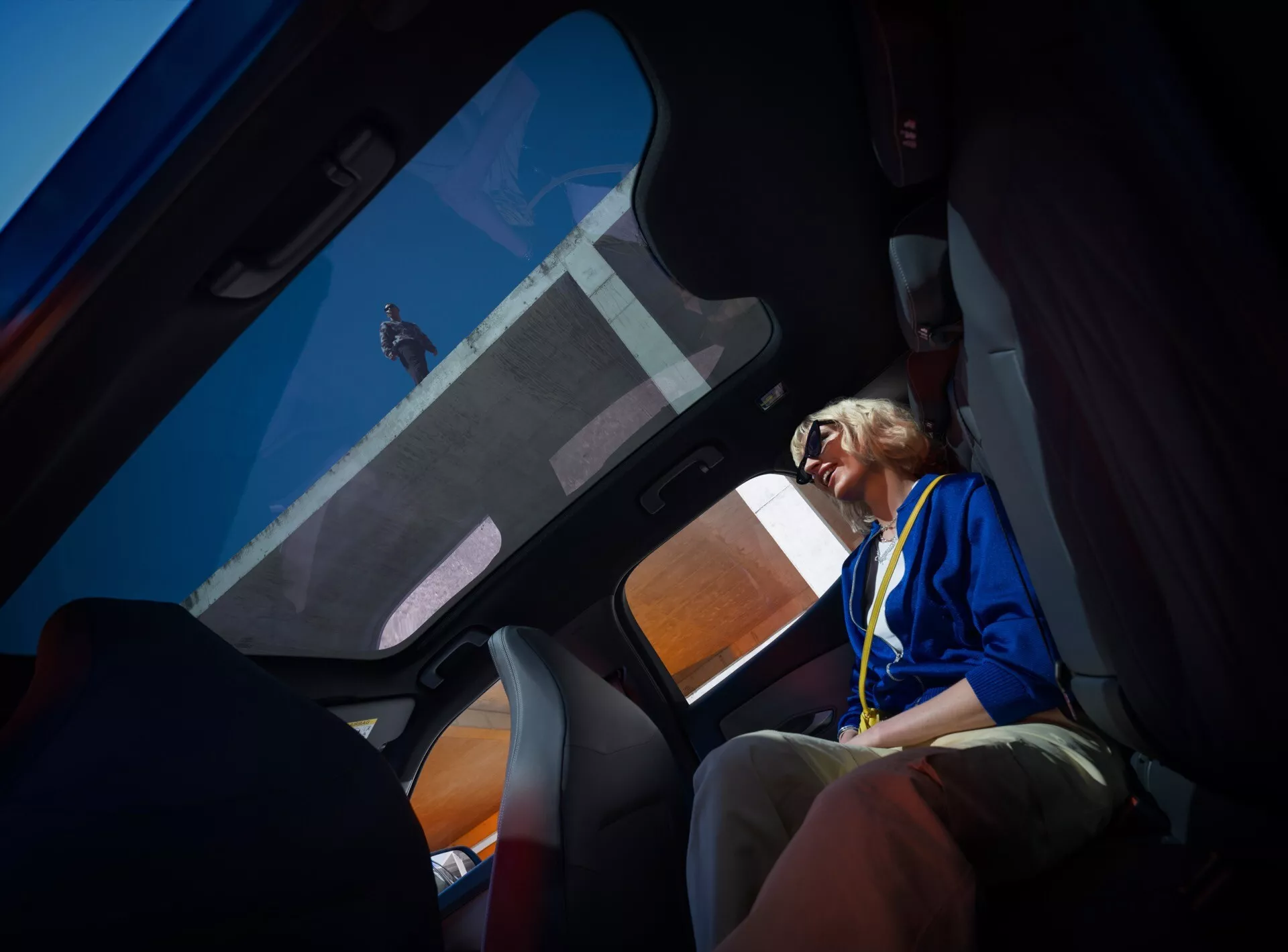 Interiér Ford Capri 2024 (4. generácia) pohľadom zo zadného sedadla, zobrazujúci panoramatické strešné okno, časť zadného sedadla, dvere spolujazdca s vysokokvalitnou úpravou a čalúnenie látkou s farebnými akcentmi.