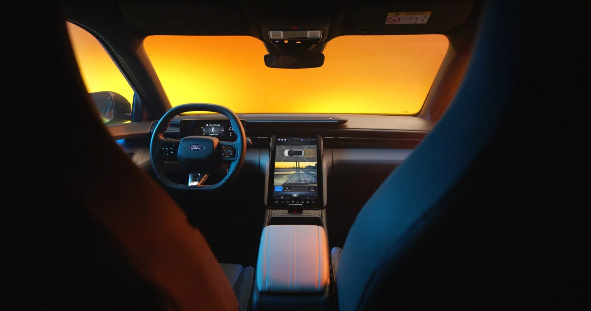 Pohľad na interiér Ford Capri 2024, ukazujúci modernú a luxusnú prístrojovú dosku a prednú kabínu z pohľadu zadného sedadla počas úsvitu alebo súmraku. Volant vľavo má multifunkčné ovládania a digitálny prístrojový panel nahrádza tradičné číselníky. Dominantou stredovej konzoly je veľký vertikálny dotykový infotainment systém, doplnený vetracími otvormi a moderným prenosovým systémom. Štýlový, minimalistický interiér zdôrazňuje pohodlie a technológiu s dvojfarebnými sedadlami pridávajúcimi estetický prvok.