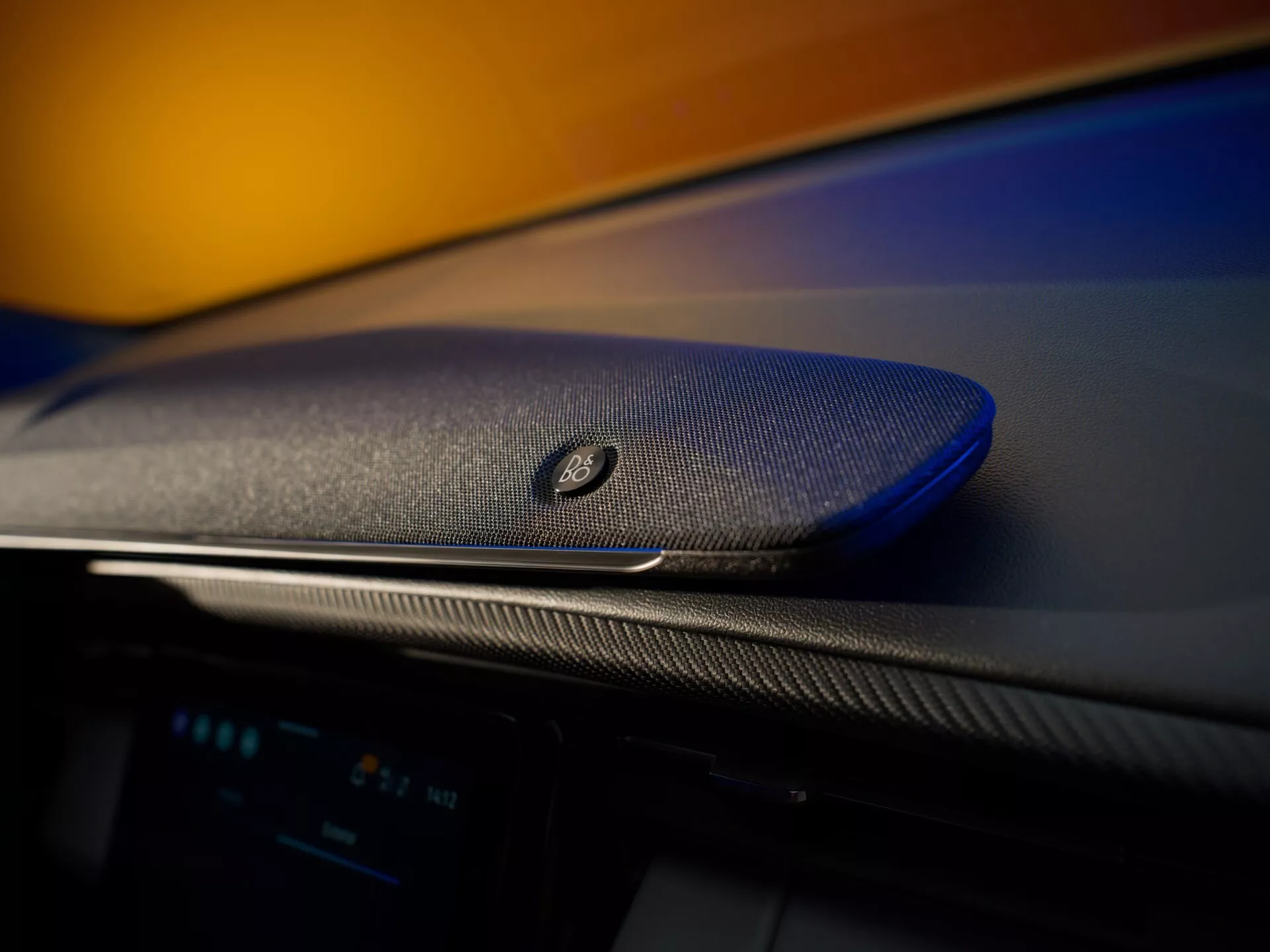 Detailný pohľad na interiér modelu Ford Capri 2024, ktorý zvýrazňuje prémiový audiosystém Bang & Olufsen, bezproblémovo integrovaný do luxusnej palubnej dosky s karbónovým povrchom. Fotografia zobrazuje textúru mriežky reproduktora v ostrom zameraní proti rozmazanému pozadiu, čím zdôrazňuje športový a elegantný vzhľad vozidla.