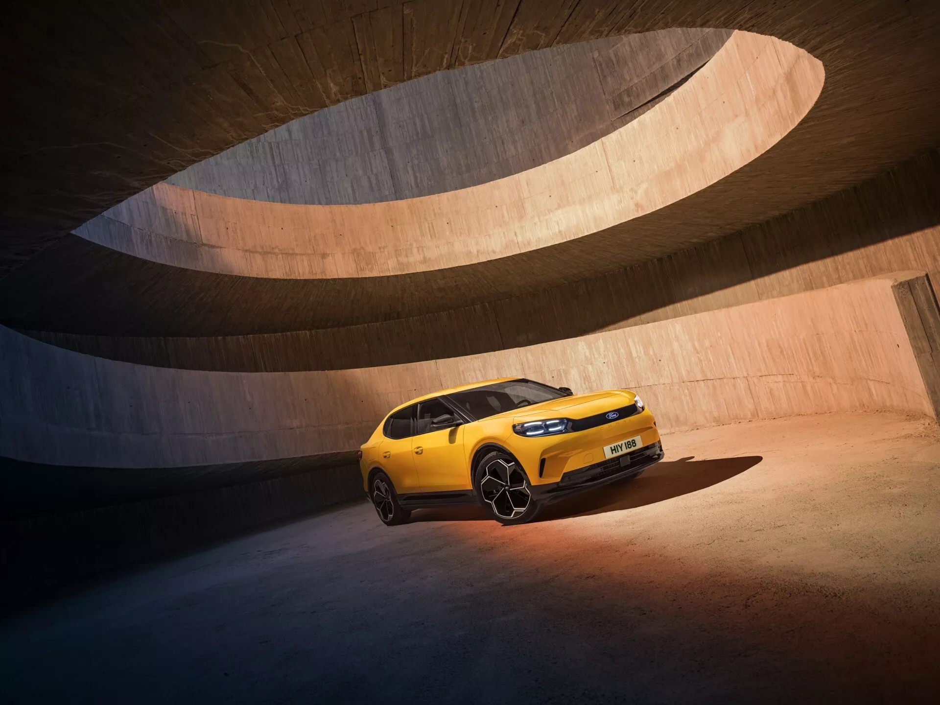 Obrázok štvrtej generácie Ford Capri 2024 zachytený z uhla troch štvrtín spredu, ukazujúci prednú časť a pasažiersky bok. Moderný hatchback alebo crossover sa vyznačuje výraznou prednou mriežkou s integrovanými LED svetlometmi, športovými nasávacími otvormi alebo ventilmi a vyššou svetlou výškou, čo naznačuje výkonovo orientované alebo terénne schopnosti. Tvarované panely karosérie a výrazné liate kolesá dodávajú dynamický postoj, zatiaľ čo jasne matná žltá farba a strešné ližiny zdôrazňujú jeho súčasný dizajn. Fotografované na pozadí zakrivených betónových štruktúr s teplým osvetlením neskorého popoludnia, obraz zdôrazňuje modernú estetiku a pokročilé funkcie vozidla.
