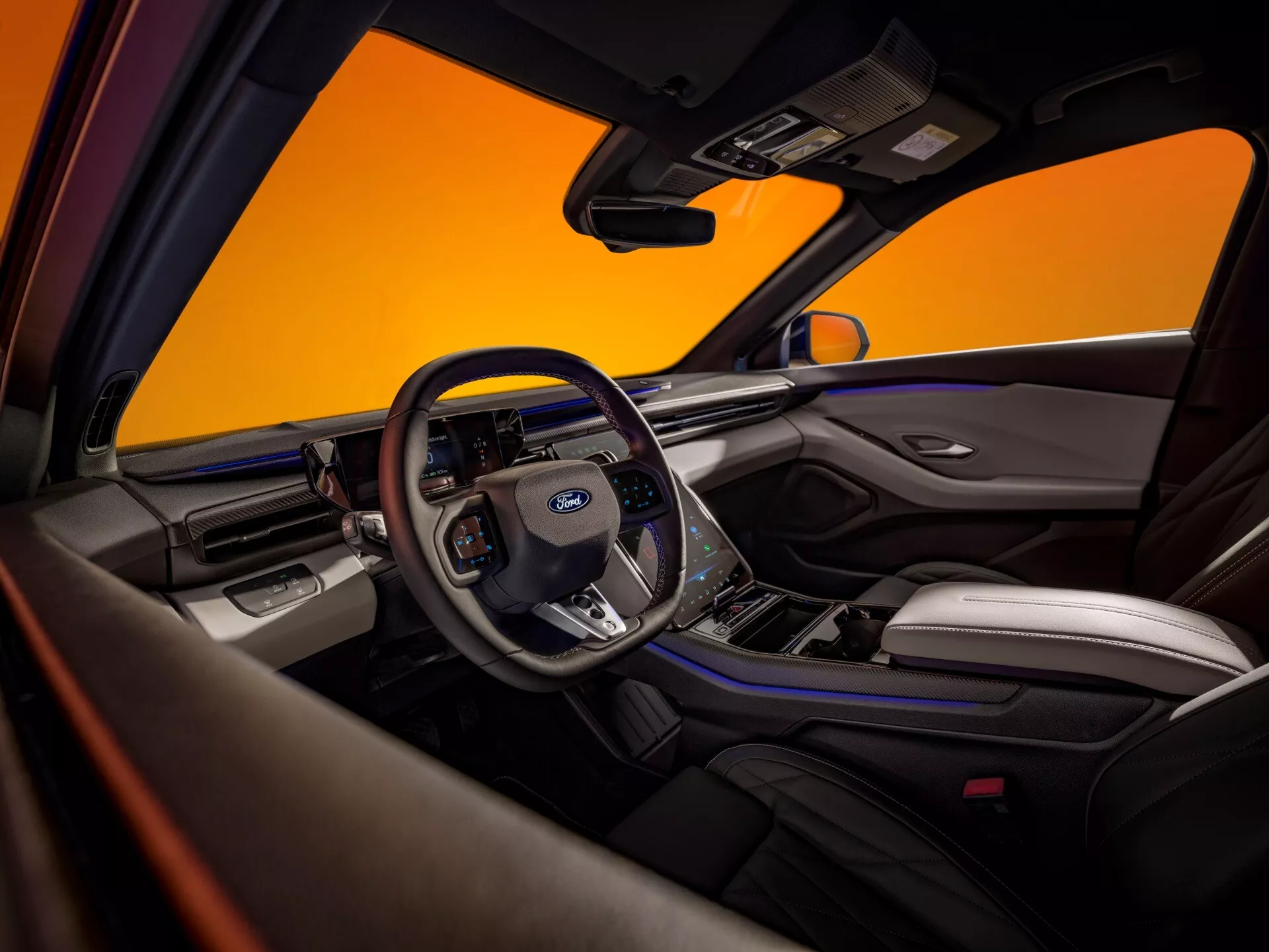 Interiér Fordu Capri 2024 štvrtej generácie, pohľad z perspektívy zadného pasažiera. Je tu výrazný volant s logom Ford s ovládacími prvkami pre audio, tempomat, hlasové príkazy a handsfree telefonovanie. Viditeľný je digitálny panel prístrojov s rýchlomerom, tachometrom a ďalšími indikátormi, spolu s centrálnym displejom infotainmentu pravdepodobne ponúkajúcim navigáciu a mediálne možnosti. Interiér ukazuje prémiovú zmes kožených sedadiel s kontrastným prešívaním, kovové a lesklé čierne povrchy. Bezpečnostné prvky zahŕňajú airbagy, bezpečnostné pásy a pokročilé asistenčné systémy vodiča. Dvojfarebná schéma interiéru zvyšuje moderný a luxusný vzhľad, zdôraznený propagačnou fotografiou s oranžovým pozadím.