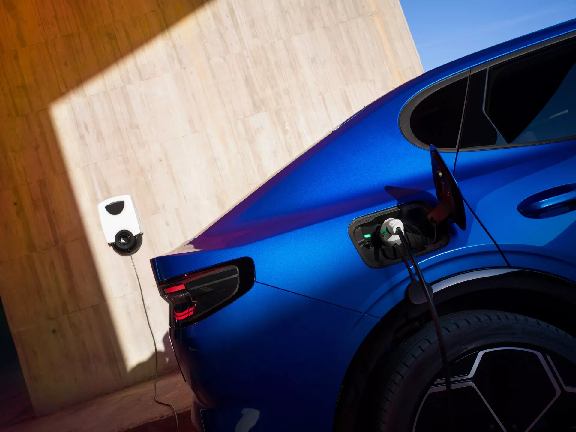 Čiastočný pohľad na modrý Ford Capri 2024 (4. generácia) nabíjajúci sa na nabíjacej stanici pre elektrické vozidlá, zachytený zo zadného trojštvrťového uhla so zameraním na pravú zadnú časť, ukazujúci výrazný dizajn zadných LED svetiel, zložité viacramenné disky a otvorený nabíjací port pripojený k káblu. Obraz zdôrazňuje športové krivky a dynamické línie auta za jasného denného svetla s rozmazaným pozadím vďaka malej hĺbke ostrosti.