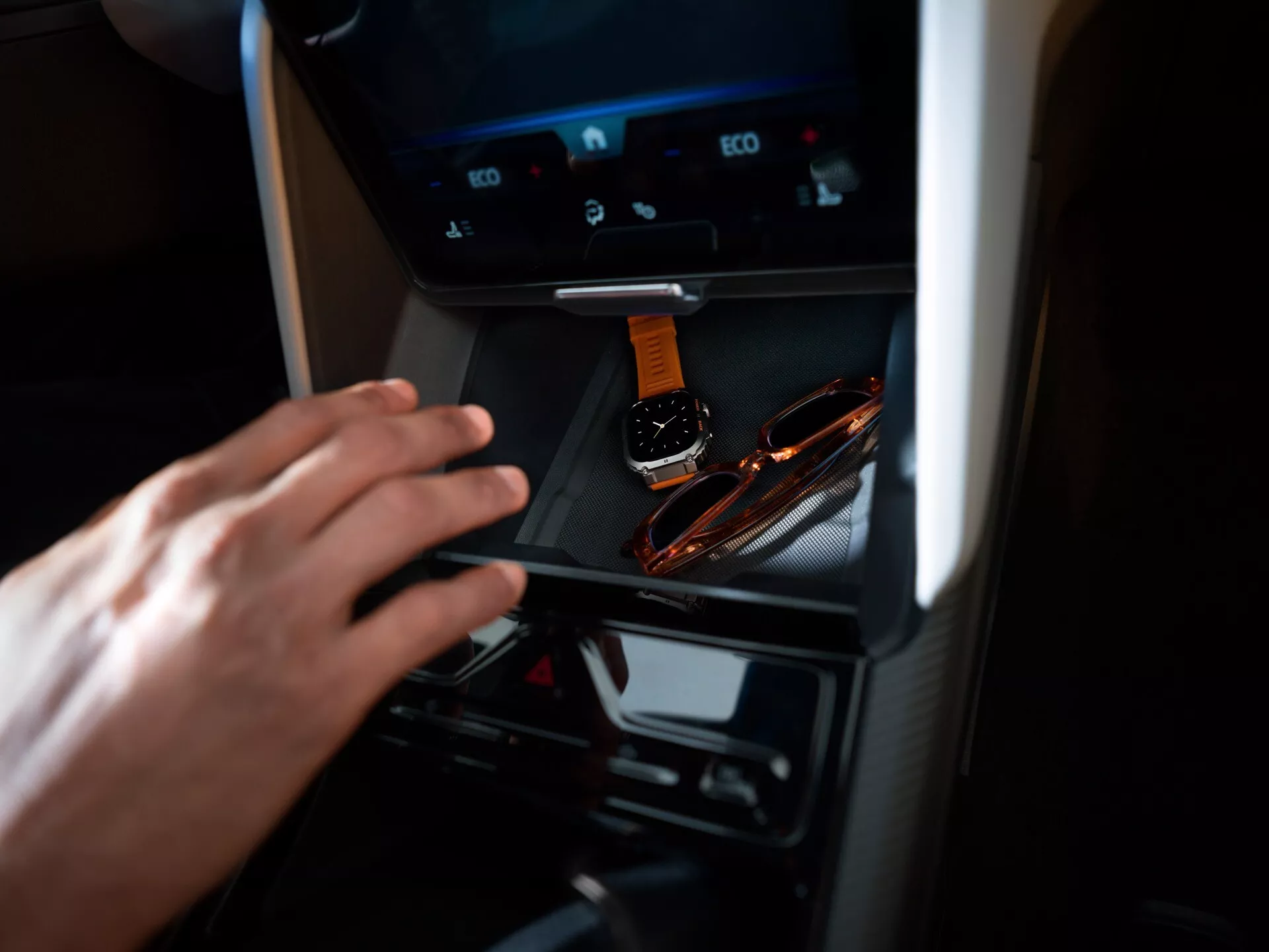 Detail interiéru Ford Capri 2024 (4. generácia) zameraný na oblasť stredovej konzoly z perspektívy spolujazdca. Obsahuje digitálny displej s ikonami klimatizácie, zabudované luxusné analógové hodiny, ruku siahať po slnečných okuliaroch v úložnom priestore a lesklú čiernu radiacu páku. Vysokokvalitná povrchová úprava s použitím kvalitných materiálov, osvetlená odrazmi slnečného žiarenia.