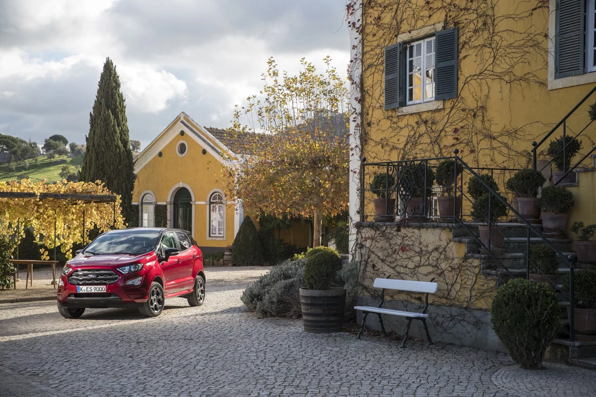 Ford EcoSport 2017 (1. generácia po facelifte) v červenej farbe, zaparkované pred okrový dom s popínavými rastlinami, pod jemným ranným alebo popoludňajším svetlom, predvádzajúce predný a bočný profil vozidla, štýlové zliatinové kolesá a moderný design LED svetlometov