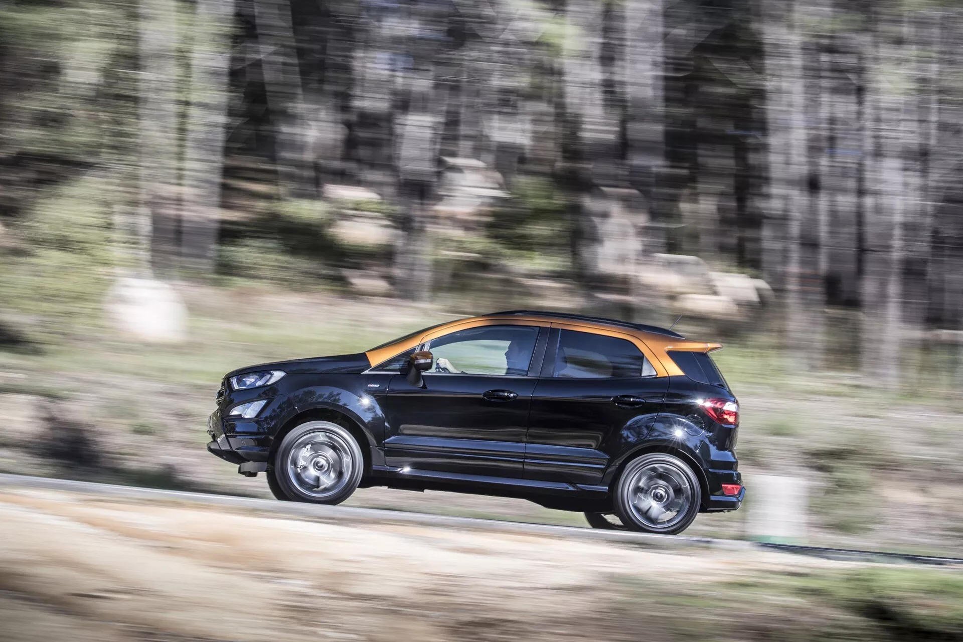 Ford EcoSport 1. generácie po facelifte z roku 2017 v dynamickom pohybe, zdôrazňujúci jeho dvojfarebné čierne a možno oranžové alebo bronzové farebné schéma, bočné a zadné dizajnové prvky, zliatinové kolesá a lesklý povrch na ceste z prachu alebo štrku, naznačujúci všestrannú schopnosť.