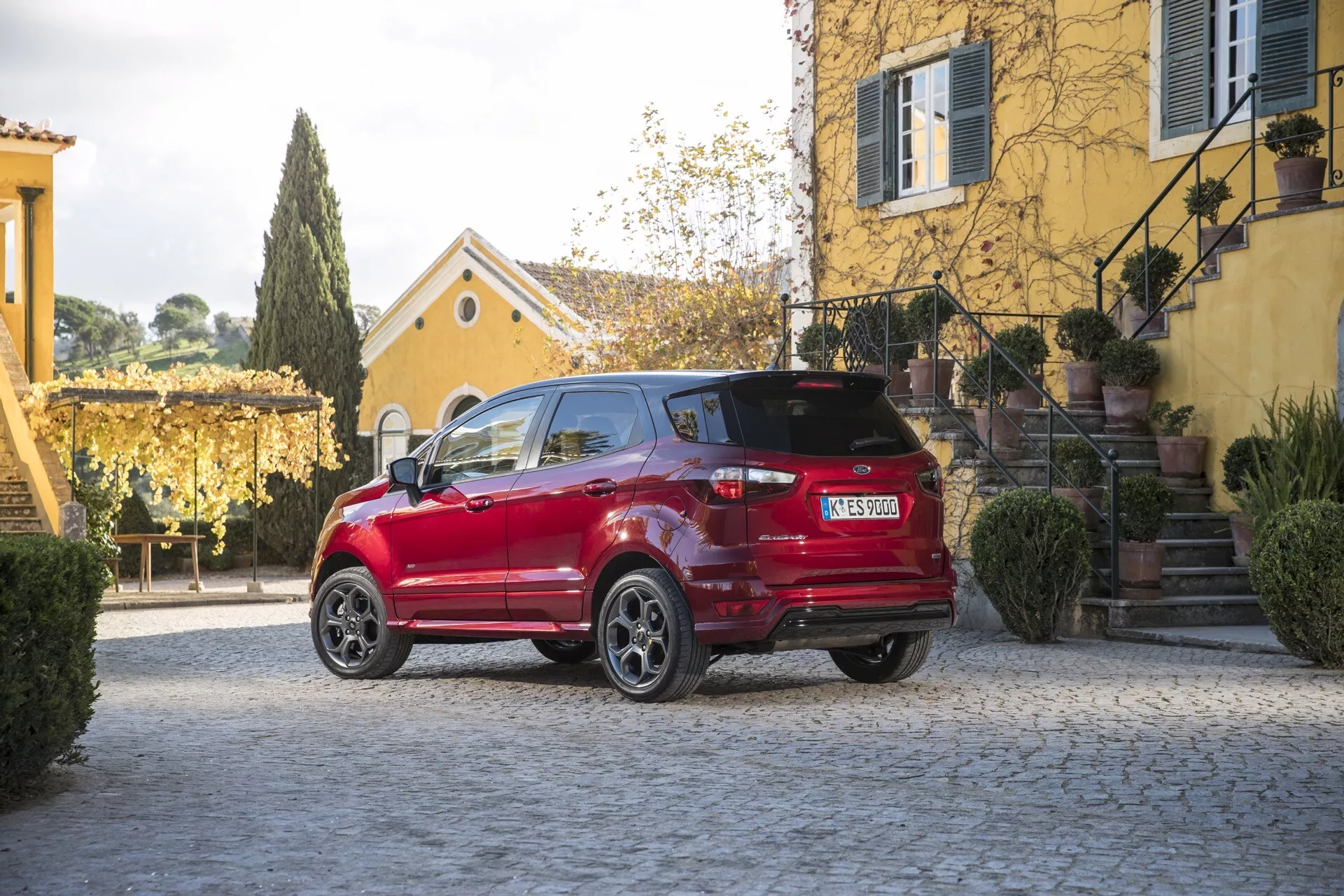 Červený Ford EcoSport 1. generácie po facelifte z roku 2017, zobrazený z trojštvrťového zadného uhla parkovaný na povrchu z dlažobných kociek s detailnými prvkami ako sú LED zadné svetlá, zliatinové kolesá, rozmazaná európska SPZ, spoiler na streche a čierne lišty okolo blatníkov viditeľné proti pokojnému rezidenčnému pozadiu s klasickou architektúrou a bujnou zeleňou v prirodzenom slnečnom svetle.