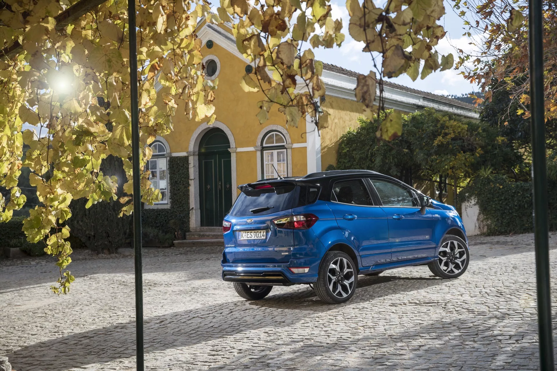 Zadný trojštvrťový pohľad na modrý Ford EcoSport (1. generácia facelift) z roku 2017, na obrázku sú viditeľné LED zadné svetlá, zadný spojler, odmrazovanie zadného skla, zadný stierač, anténa v tvare žraločej plutvy, senzory pre parkovanie, dvojité výfuky a zliatinové disky s viacerými ramenami, zaparkovaný na dlažobných kockách pri žltej vile so zelenými okenicami.