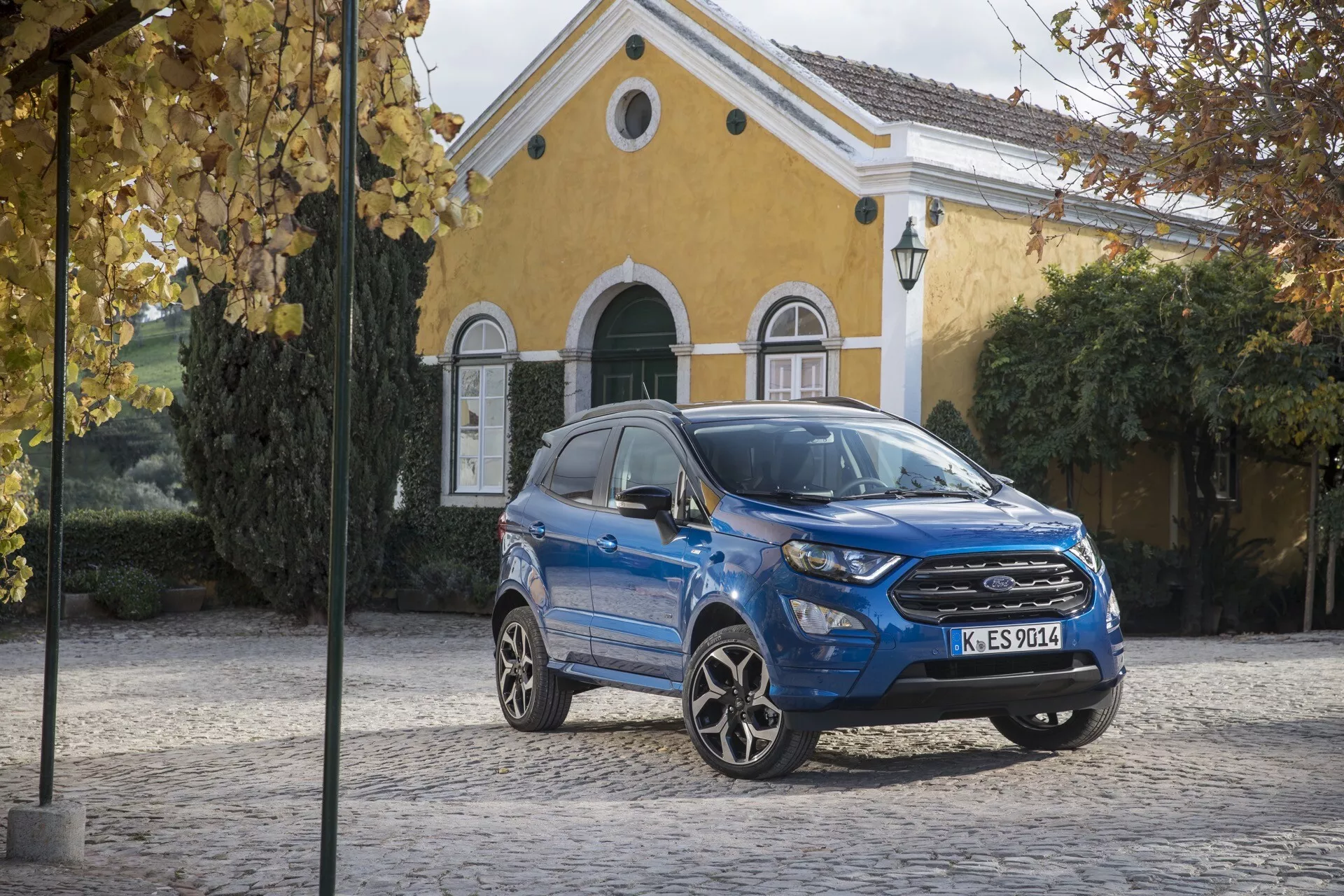 Modré kompaktné SUV Ford EcoSport Facelift 2017 parkované na dlažobnom kameni so žltou budovou v pozadí, prezentujúce prednú mriežku, LED svetlomety a športové zliatinové kolesá