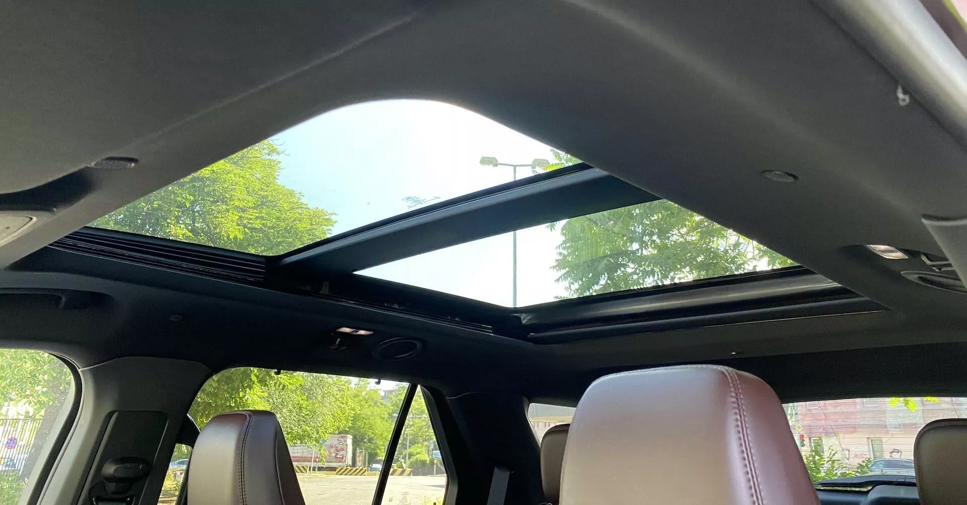 Interiérový pohľad na Ford Explorer 2019 so zameraním na panoramatické strešné okno z priestoru zadných sedadiel. Strešné okno sa tiahne od prednej časti dozadu s veľkým skleneným panelom, ktorý umožňuje dostatok prirodzeného svetla, doplnené otvorenou zasúvacou slnečnou clonou. Predná časť strechy obsahuje ovládacie prvky a čítacie svetlo na stropnej konzole. Tmavý stropný obklad kontrastuje so svetlými koženými sedadlami, čím ukazuje moderný dizajn a luxusné prvky interiéru.