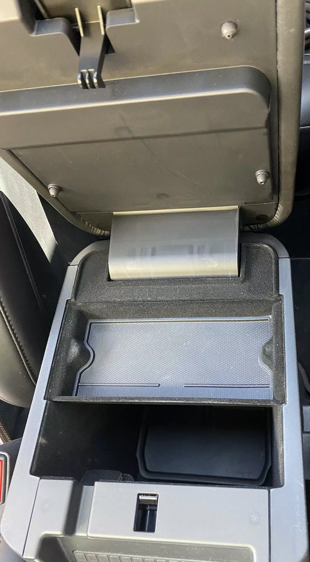 Detail interiéru Ford Explorer 2019 zameriavajúci sa na stredovú konzolu zobrazujúcu otvorenú stredovú opierku s posuvným sekundárnym zásobníkom zasunutým vzadu, odkrývajúci USB port a hlbší úložný priestor v čiernom plaste.