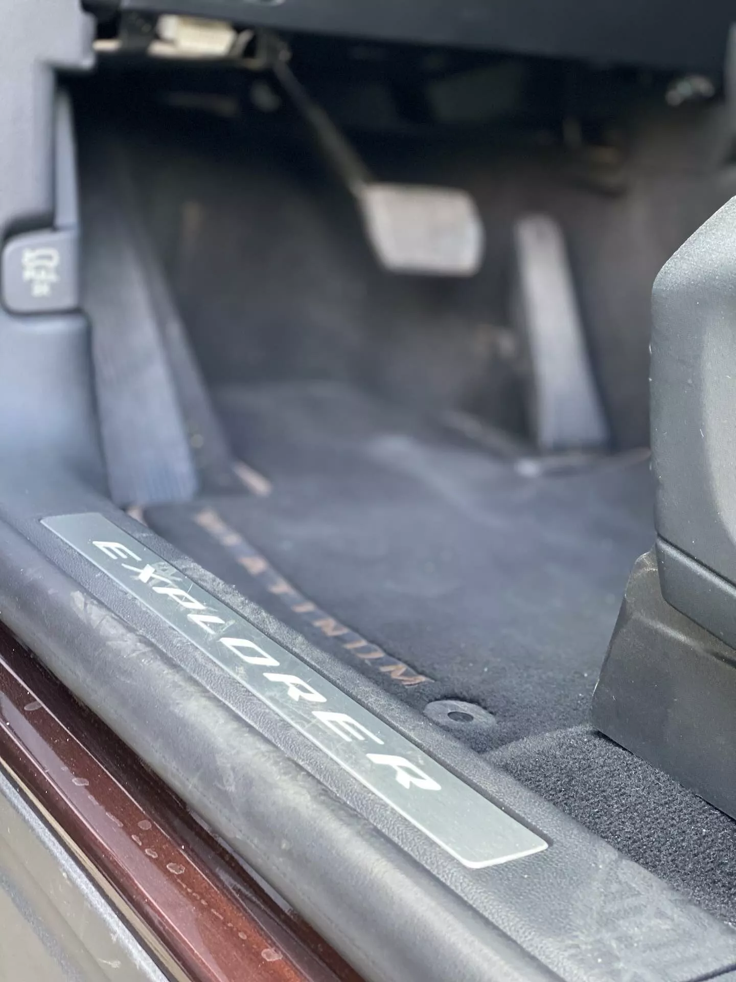 Detailný záber interiéru SUV Ford Explorer z roku 2019, ktorý konkrétne ukazuje oblasť prahu dverí s kartáčovanou kovovou doskou s vyrytým logom 'Explorer'. Obrázok zdôrazňuje prah, kde nastupujú pasažieri, s tmavým kobercom, tesnením a mäkkým prirodzeným osvetlením.