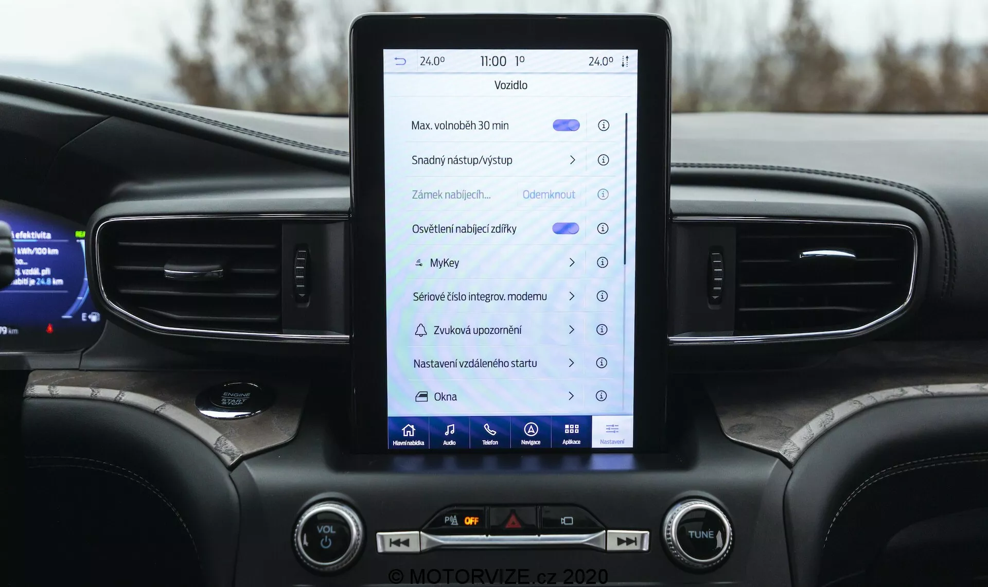 Detailný pohľad na interiér modelu Ford Explorer 2019, zameraný na stredovú konzolu a infotainment systém. Vertikálne orientovaný dotykový displej zobrazuje ikony a texty, ktoré naznačujú funkcie ako navigácia, nastavenia vozidla, ovládanie médií, Bluetooth a možno Apple CarPlay alebo Android Auto. Palubná doska zahrnuje vertikálne vetracie otvory, ambientné osvetlenie, fyzické tlačidlá a gombíky na ovládanie klimatizácie a hlasitosti. Tiež je viditeľná horná časť multifunkčného volantu a čiastočný digitálny prístrojový panel zobrazujúci informácie o vozidle. Dizajn podčiarkuje moderné vozidlo strednej alebo vyššej triedy s tmavou farebnou schémou.