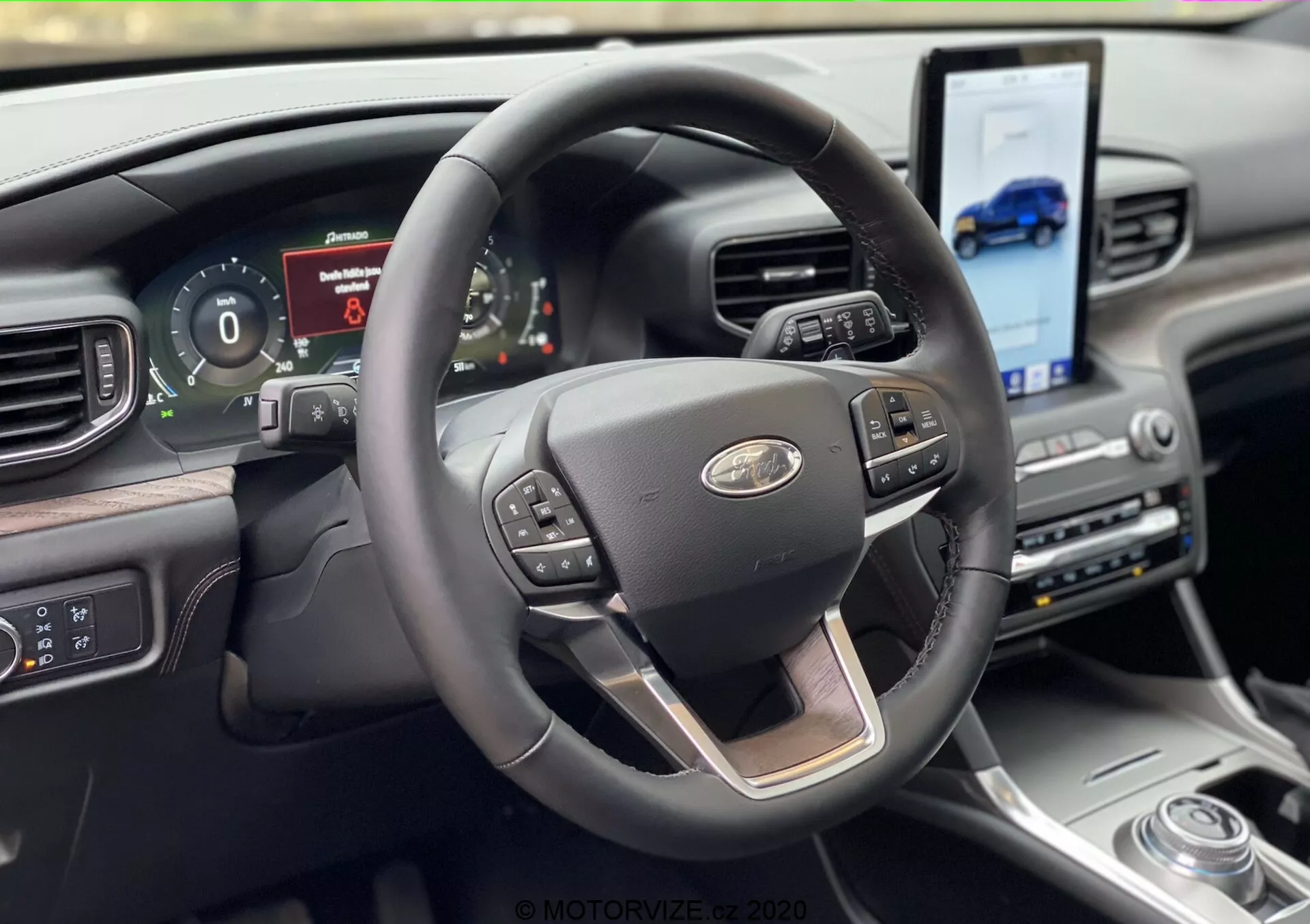 Pohľad do interiéru vozidla Ford Explorer 2019 z perspektívy sedadla vodiča, s moderným trojramenným volantom s chrómovým akcentom a multifunkčnými ovládacími prvkami pre tempomat, médiá a hlasové ovládanie. Za volantom sa nachádza digitálny prístrojový panel zobrazujúci informácie o vozidle. Palubná doska obsahuje veľký dotykový displej naklonený k vodičovi pre navigáciu a prístup k médiám, doplnený otočným voličom prevodových stupňov a luxusnými materiálmi.