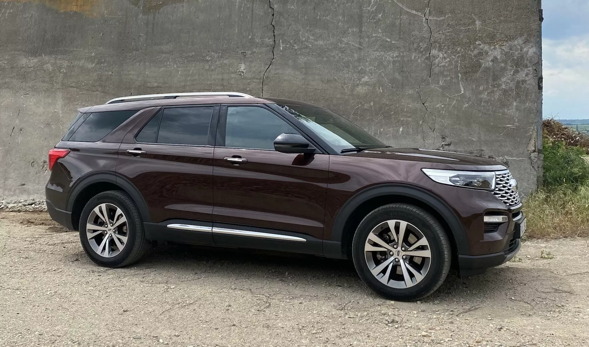 SUV Ford Explorer z roku 2019 zaparkovaný na hrubej štrkovej ploche v blízkosti betónovej štruktúry, bočný profil zvýrazňujúci dizajnové prvky ako veľké zliatinové kolesá, vysokú bočnú líniu, tónované okná, kľučky dverí vo farbe karosérie, chrómový prúžok na oknách, elegantné línie karosérie, zrkadlá so zabudovanými smerovkami, hlboký metalický lak a znateľnú svetlú výšku.
