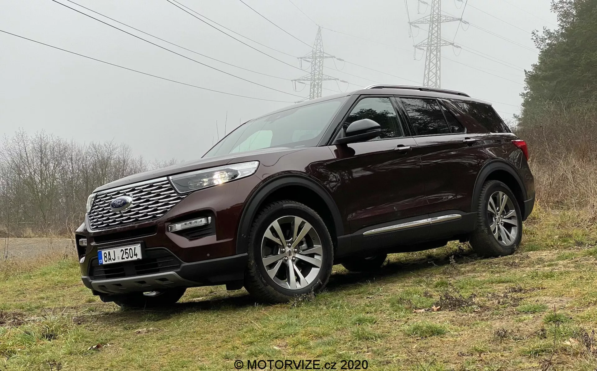 Obrázok tmavého SUV Ford Explorer 2019 zaparkovaného na nerovnom trávnatom teréne, zdôrazňujúci jeho robustnosť a schopnosť jazdy v teréne. Zachytené z predného trojštvrťového uhla, SUV jasne ukazuje dizajnové prvky ako výraznú mriežku s geometrickým vzorom, emblém značky, široký prieduch, hmlovky, elegantné obalujúce LED svetlomety s kovovými akcentmi a viacramenné zliatinové kolesá. Funkcie zahŕňajú aj zrkadlá vo farbe karosérie s integrovanými smerovkami a strešnými nosičmi, čo naznačuje praktickosť a sofistikovanosť.