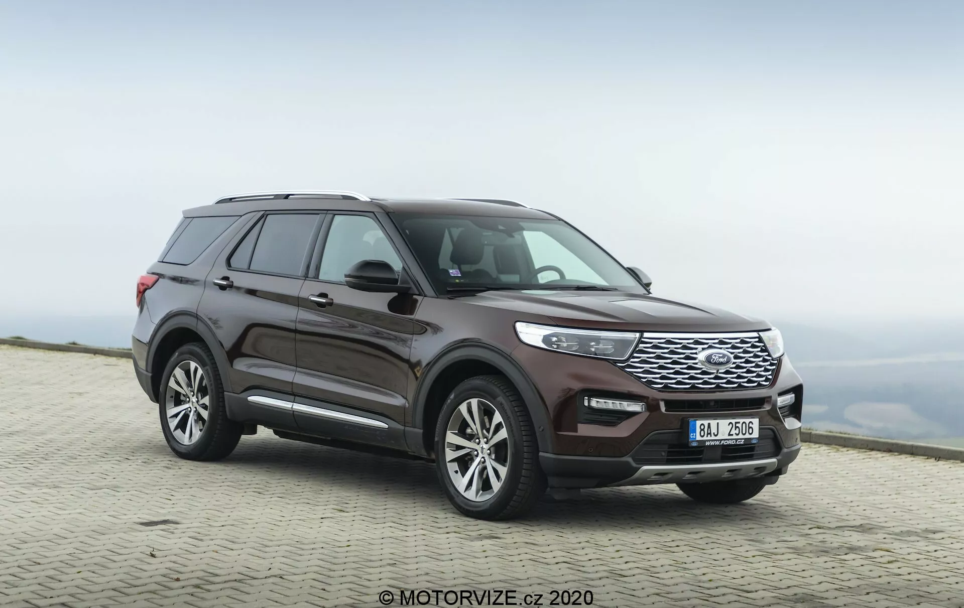 Obrázok SUV Ford Explorer 2019 zachyteného z trojštvrťového predného uhla, zvýrazňujúc bočný profil a prednú časť vozidla. Nízky uhol kamery zdôrazňuje robustný postoj a výraznú prednú mriežku vozidla. Celá dĺžka vozidla je viditeľná, ukazujúc dizajnové línie, dizajn kolies a kontúry dverí a blatníkov. Kolesá sú mierne otočené doprava, čo dodáva pocit pohybu. Výrazné prvky zahŕňajú prednú mriežku s vodorovnými lamelami, elegantné svetlomety, ktoré sa integrujú s mriežkou, a predný nárazník s viditeľnými senzormi. Bočné zrkadlá pravdepodobne obsahujú integrované smerovky a strešná línia sa mierne zvažuje dozadu, s viditeľnými strešnými koľajnicami. Farba povrchu je hlboká kovová burgundská. Vozidlo je zaparkované na dláždenom povrchu s rozmazaným prírodným pozadím, čo naznačuje vysokú nadmorskú výšku s oblačným nebom.