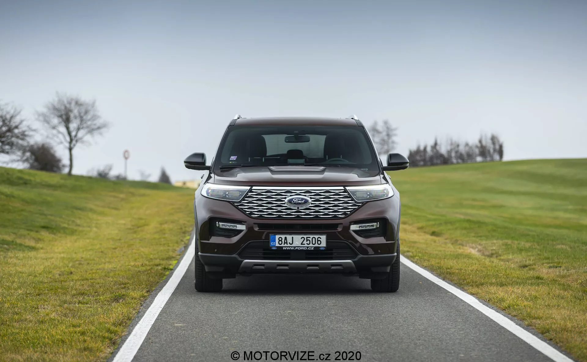 Čelný pohľad na moderné auto, ktoré zaberá stredový rám na dvojprúdovej ceste, s elegantným dizajnom svetlometov s dennými LED svetlami, výraznou prednou mriežkou so vzorom včelieho plástu a pokročilými aerodynamickými obrysmi na nárazníku, pravdepodobne obsahujúcimi parkovacie senzory a ďalšie svetlá.