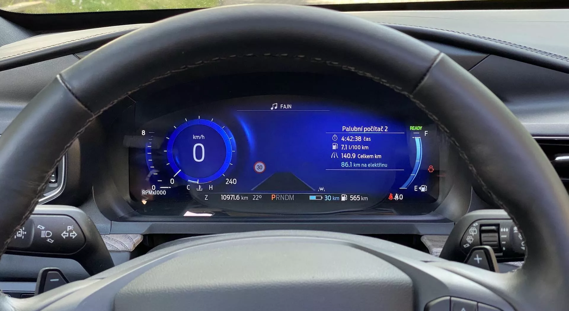 Interiér auta Ford Explorer 2019 z pohľadu vodiča, zameraný na centrálne umiestnený volant a digitálnu prístrojovú dosku za ním. Volant má rôzne ovládacie tlačidlá na oboch ramenách. Za ním je plne digitálny prístrojový panel, ktorý zobrazuje informácie ako rýchlosť, ukazovateľ výkonu, dojazd, úroveň paliva, vonkajšiu teplotu a údaj o spotrebe. Prístrojová doska zobrazuje aj čas a dátum a je navrhnutá s modernými, technologicky pokročilými prvkami v modrej a bielej farbe. Volant je vyrobený z čiernej kože so švami.