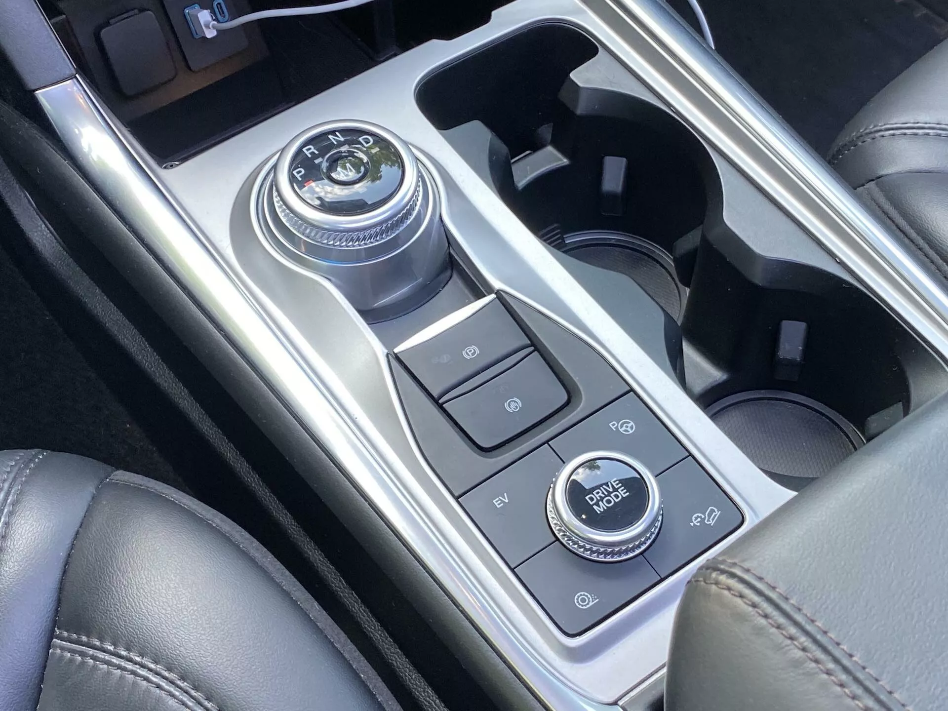 Detailný pohľad na stredovú konzolu vozidla Ford Explorer 2019 s rotačným voličom prevodovky, rôznymi ovládacími tlačidlami, držiakmi na nápoje a ovládacím kolieskom infotainment systému, všetko obklopené lesklými kovovými a matnými úsekmi, ktoré naznačujú moderný luxus a pokročilé technológie.