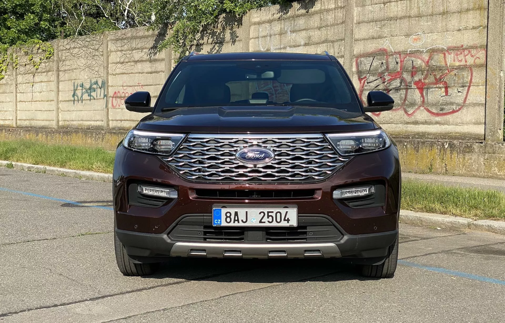 Čelný pohľad na SUV Ford Explorer 2019 zachytený z úrovňového uhla, ktorý zvýrazňuje moderný dizajn vozidla. Výrazná predná časť vozidla obsahuje veľkú chrómovanú mriežku so šesťuholníkovým vzorom a centrálnym znakom výrobcu. Štýlové, horizontálne predĺžené LED svetlá a nárazník vo farbe vozidla s dolnými prívodmi vzduchu a parkovacími senzormi zdôrazňujú technologické pokroky. Kapota ukazuje tvarovaný dizajn so zvýšenými líniami, zatiaľ čo blatníky a spätné zrkadlá s integrovanými smerovkami dodávajú svalnatý vzhľad. SUV, natreté bohatou gaštanovou alebo tmavočervenou farbou, je zaparkované na dláždenej ploche s betónovým obrubníkom a graffiti stenou v pozadí, čo zdôrazňuje mestské prostredie.