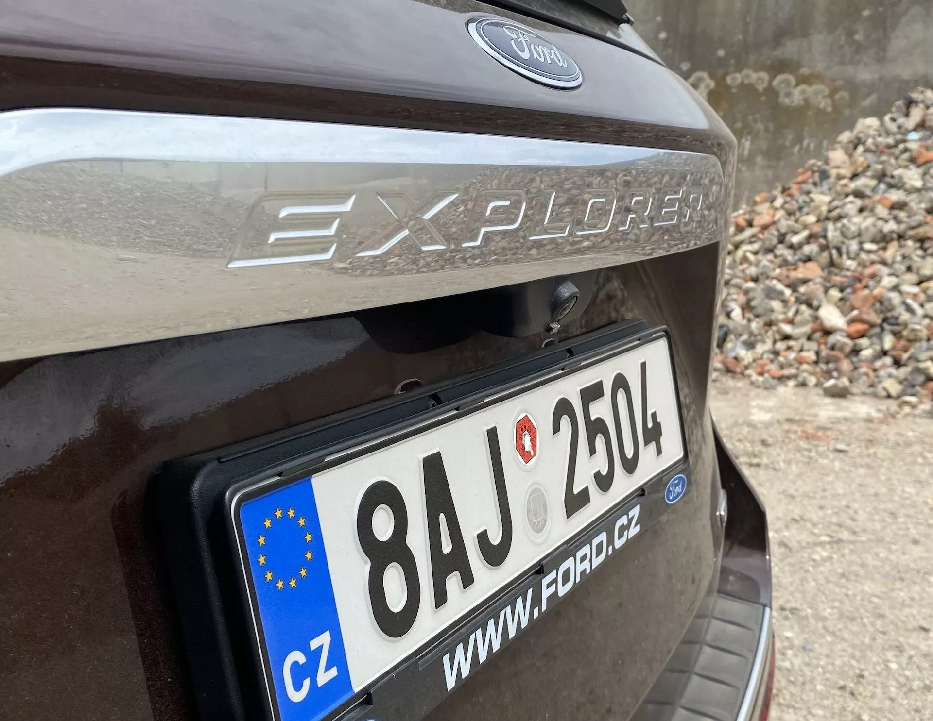 Detailný pohľad na zadnú časť vozidla Ford Explorer 2019, zameraný na oblasť zadných dverí s výrazne zobrazenou európskou registračnou značkou '8AJ 2504' a krajinským kódom 'CZ'. Názov modelu 'Explorer' a logo Ford sú nad značkou, nad registračnou značkou je viditeľná reflexná páska. Auto je pravdepodobne tmavohnedé alebo tmavošedé, zaparkované v oblasti s kameňmi alebo sutinou.